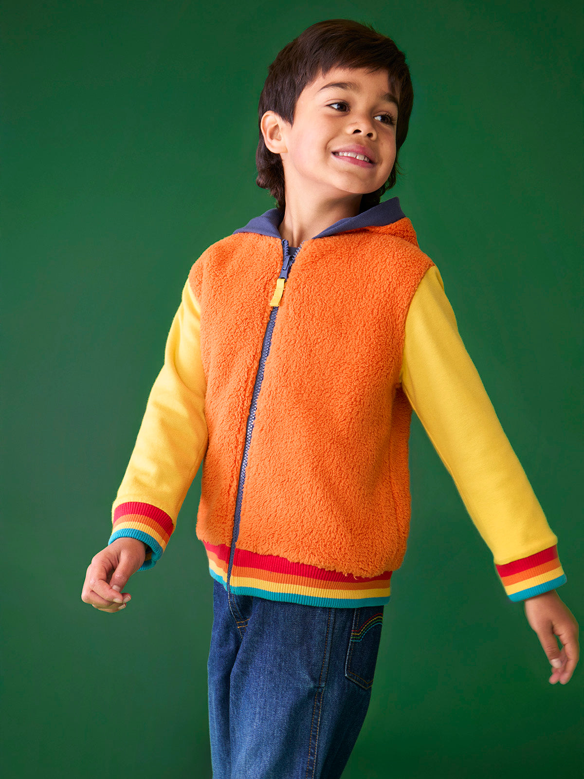 A young boy smiling while wearing an orange and yellow Frugi organic cotton hoody against a green background.