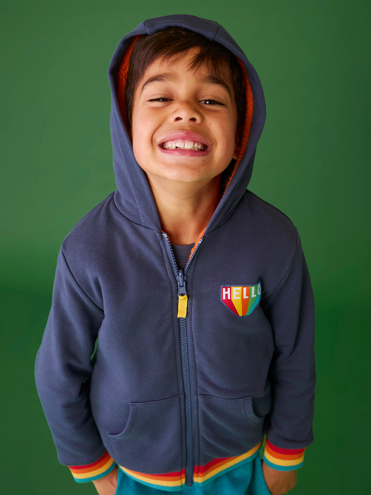 A happy young boy wearing a navy blue Frugi organic cotton hoody with a colorful rainbow Hello patch.