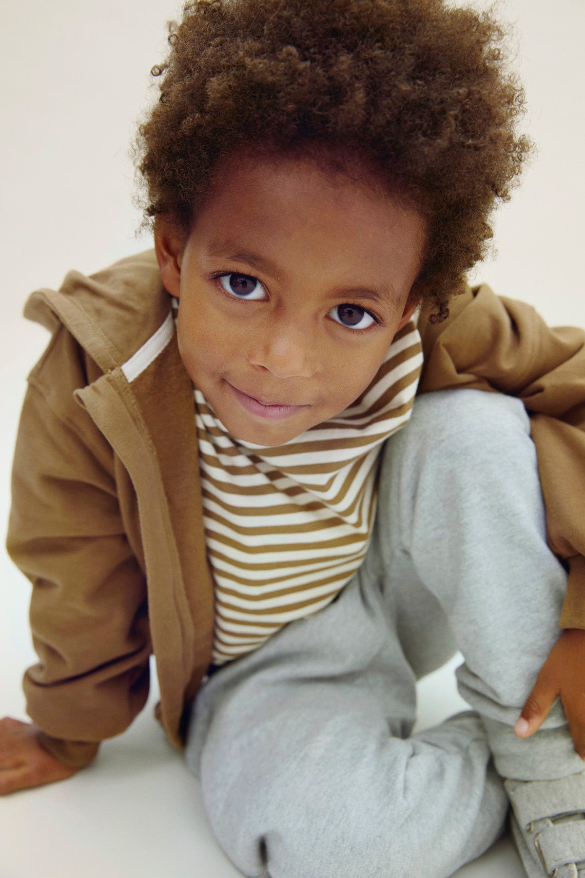 Child wearing a brown jacket, striped shirt, and gray pants on a white background