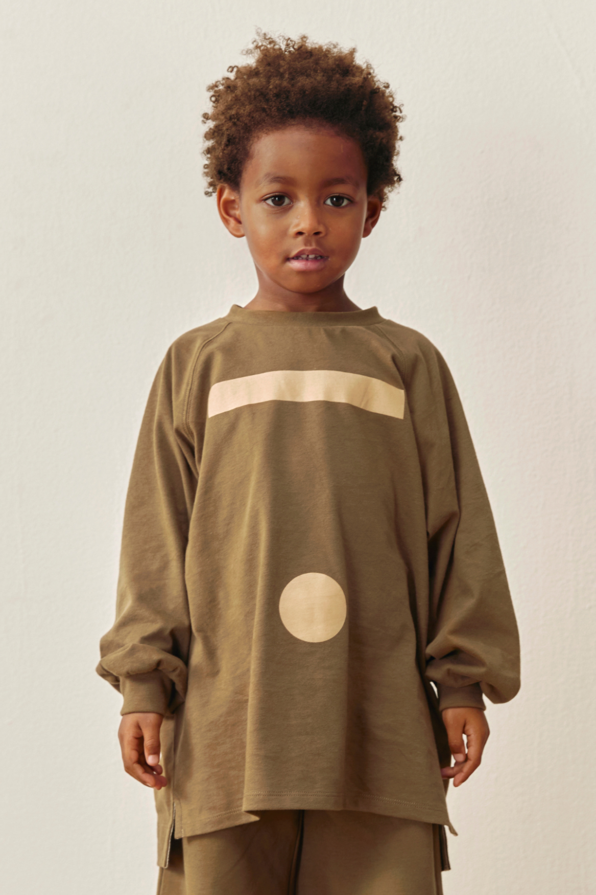 A young child with curly brown hair wearing a loose-fitting, long-sleeve olive-brown sweatshirt with a cream horizontal stripe across the chest and a matching cream circle graphic, paired with matching brown pants, against a plain off-white background.