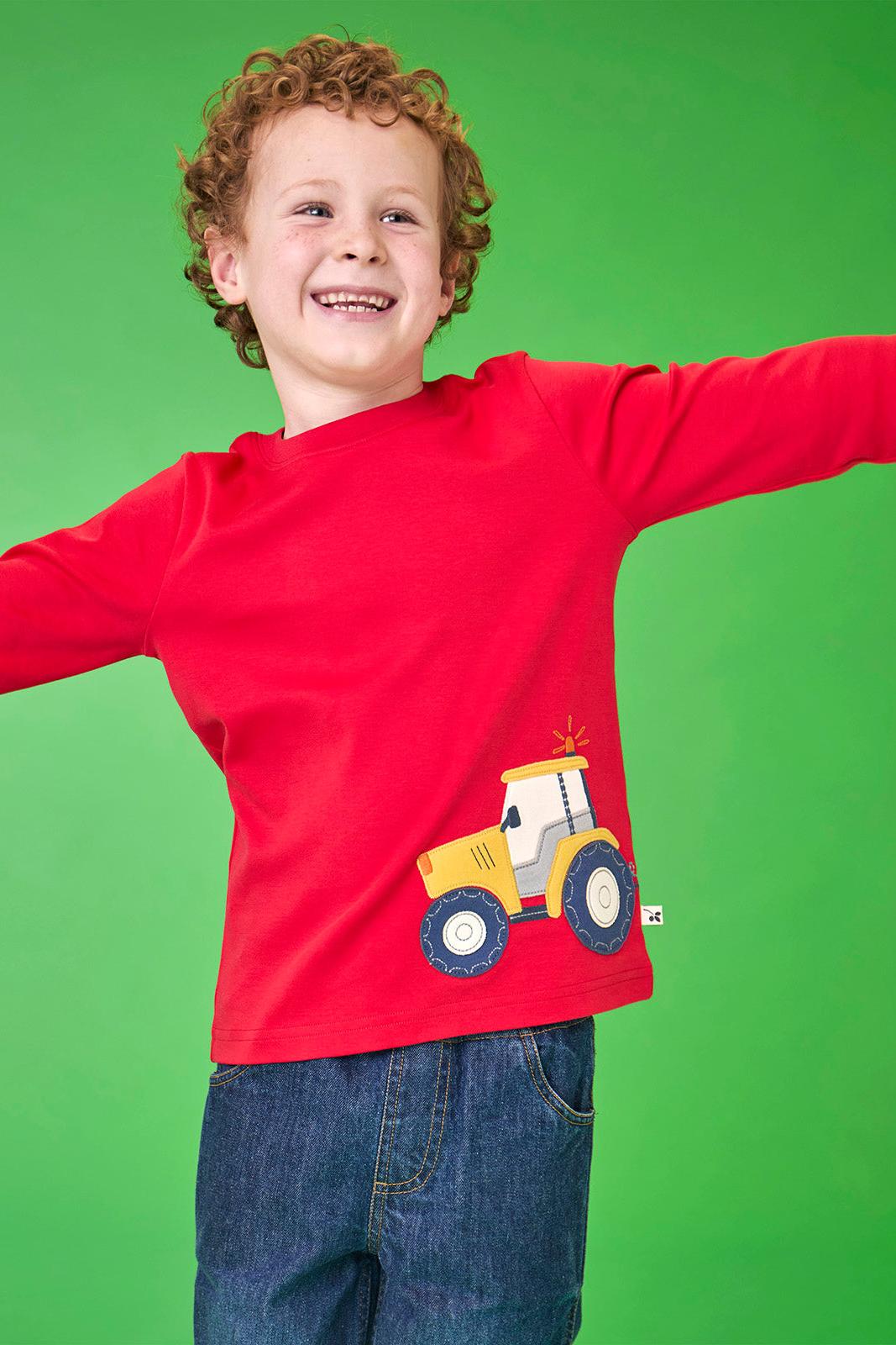 A happy young boy wearing a red organic cotton Frugi tshirt featuring a yellow tractor design.