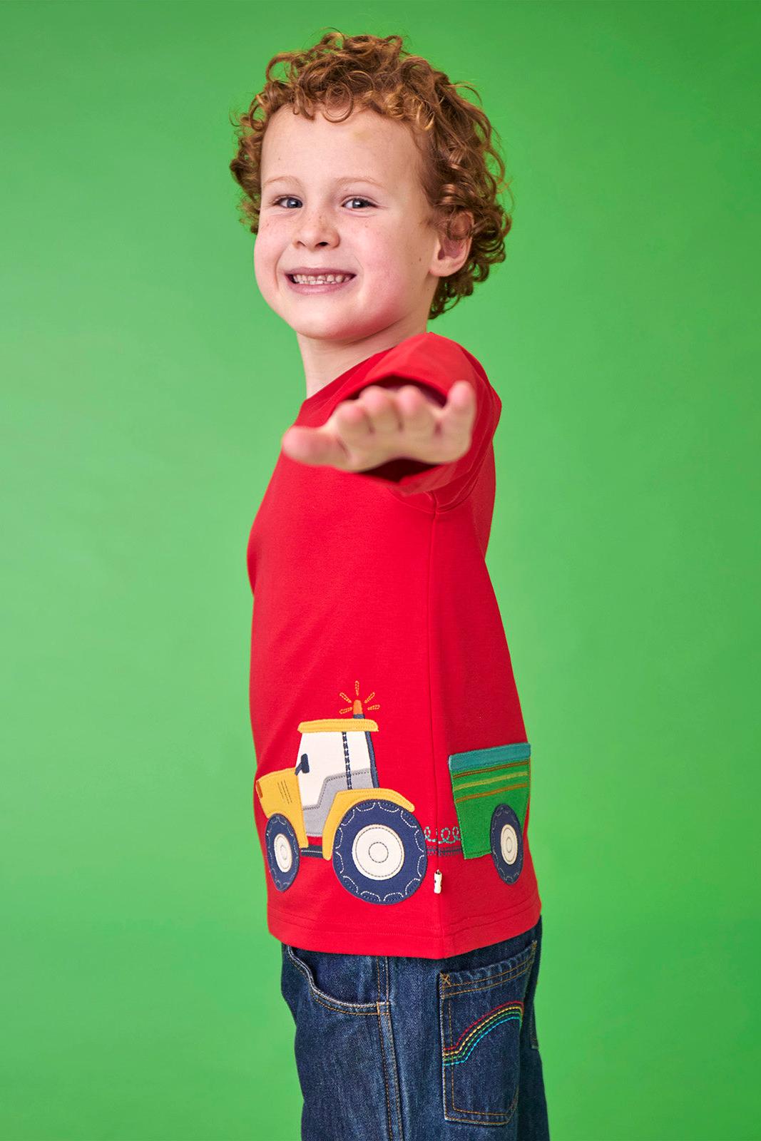A young boy with curly hair smiling while wearing a red Frugi organic cotton t-shirt with a tractor print.