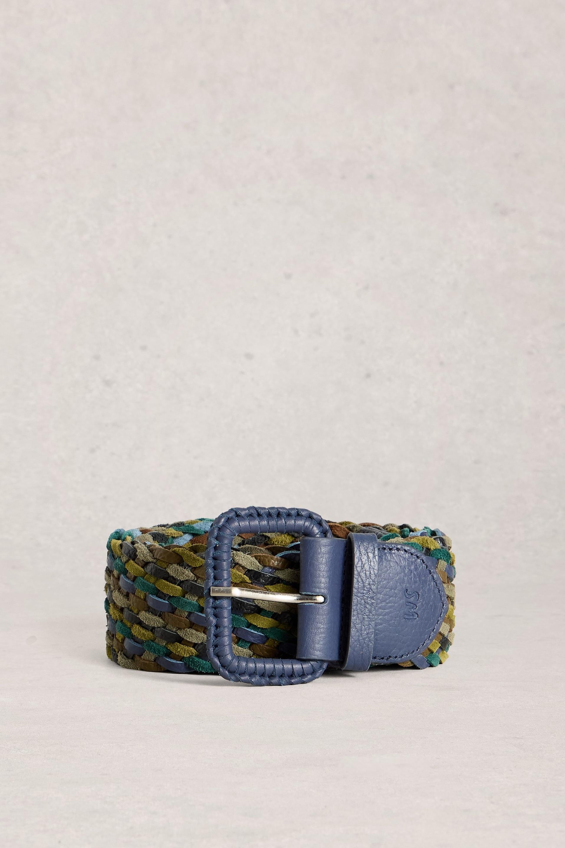 A woven belt with a blue leather buckle and keeper, featuring a multicolor braided strap in shades of green, blue, and gold, set against a neutral textured background.