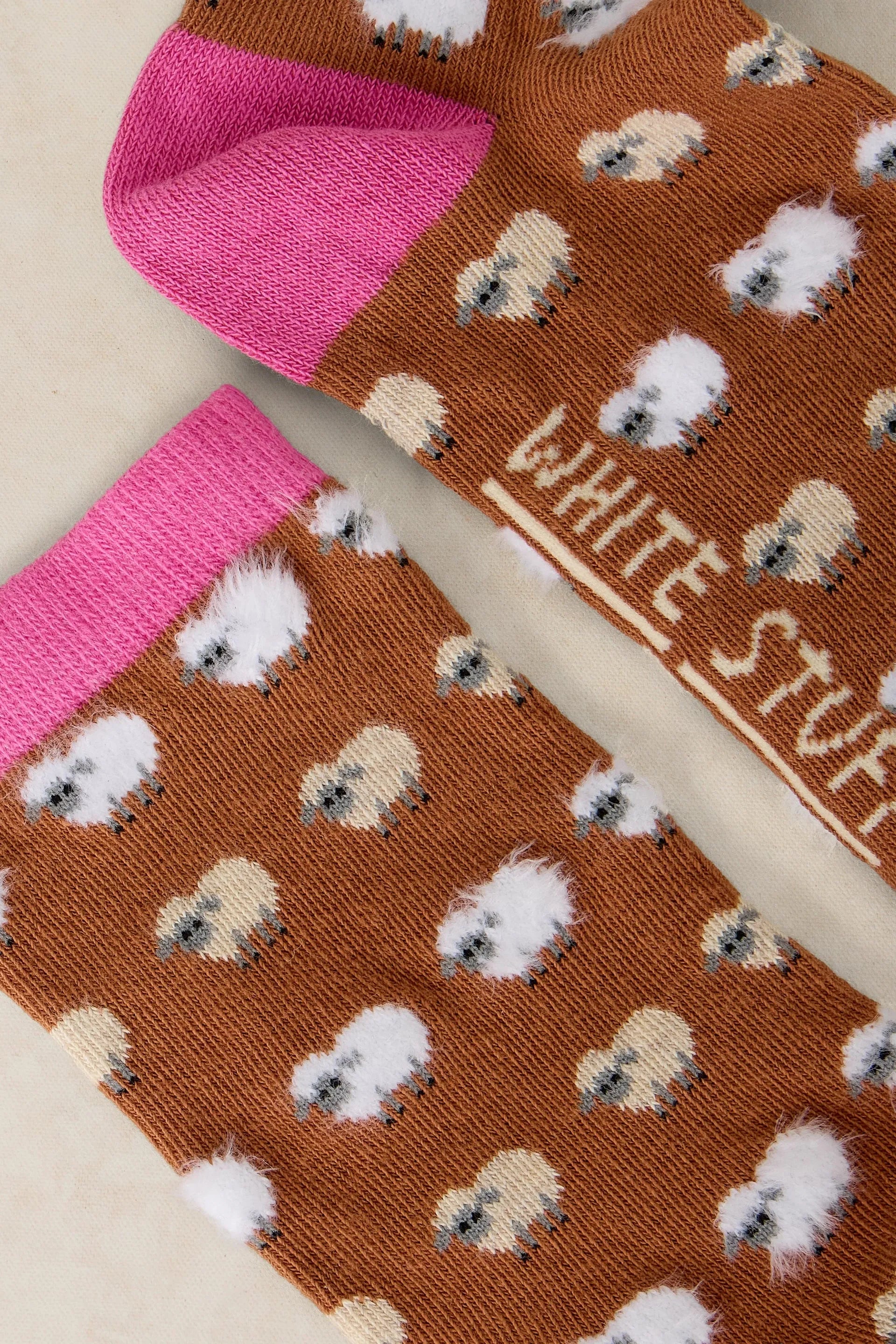 A close-up of a pair of brown socks with a repeating white sheep print, bright pink heels and cuffs, and the words 'WHITE STUFF' printed on the side.