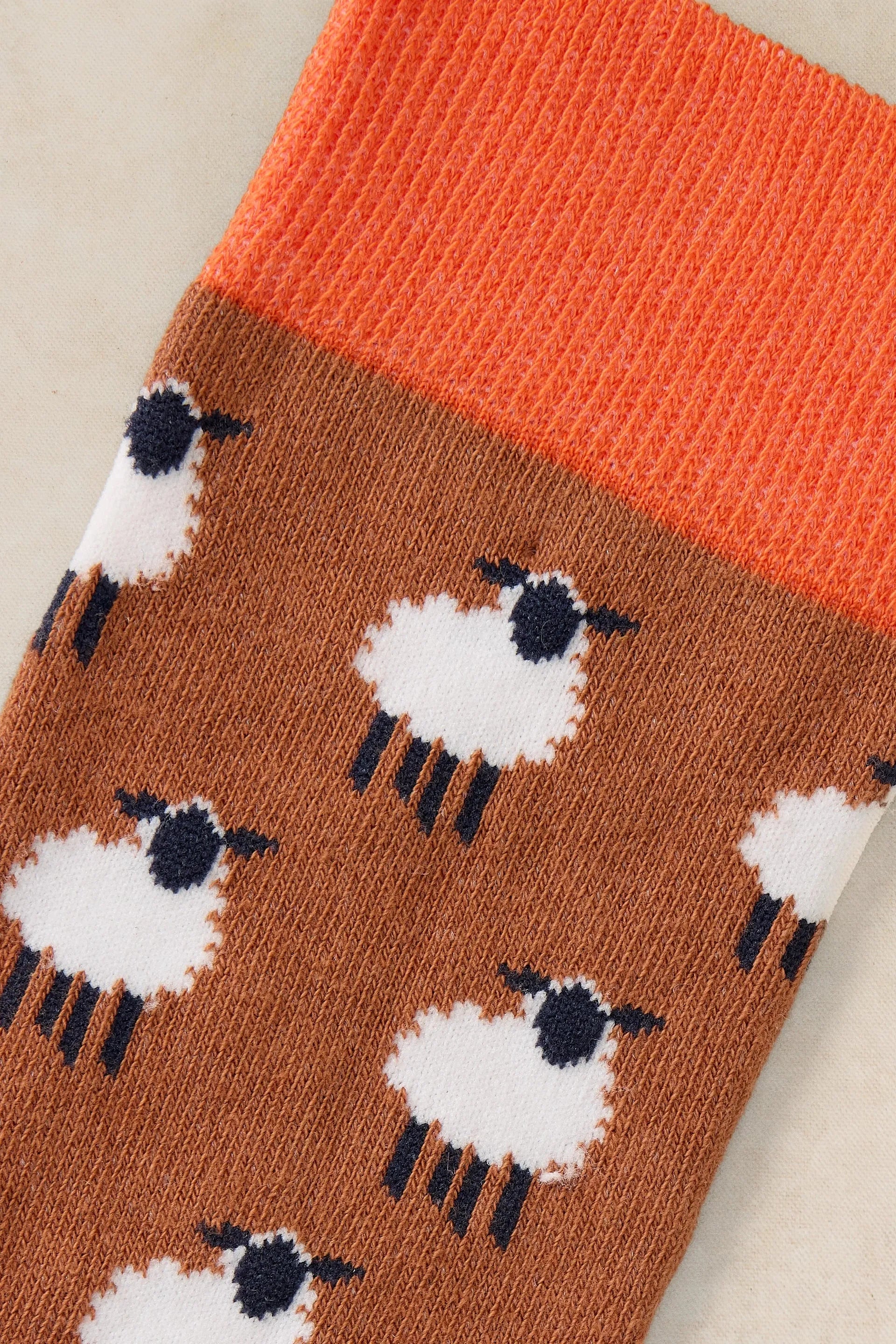 A close-up of a knit sock featuring a pattern of white sheep with black faces and legs on a warm brown background, with a bright orange ribbed cuff.