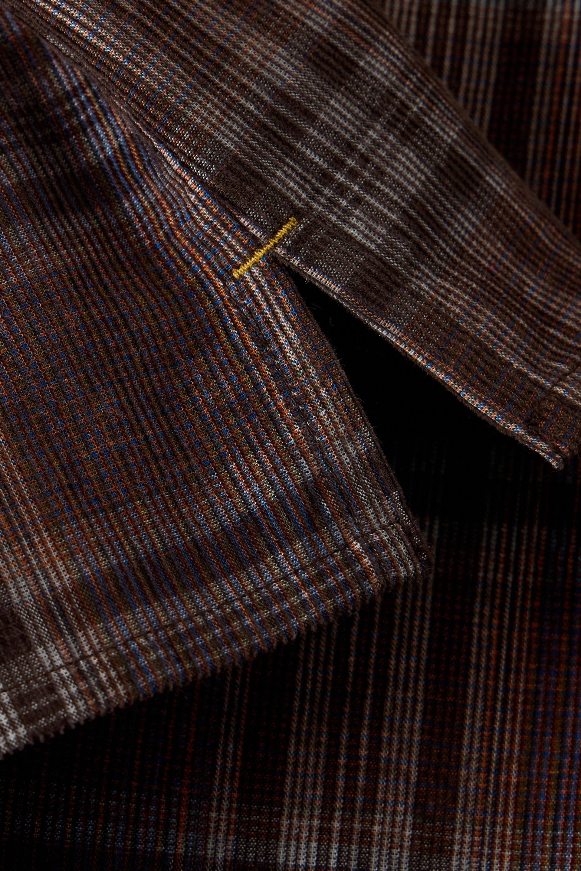 Close-up of a dark brown plaid fabric with fine blue, orange, and white stripes, showing a small yellow stitched detail.