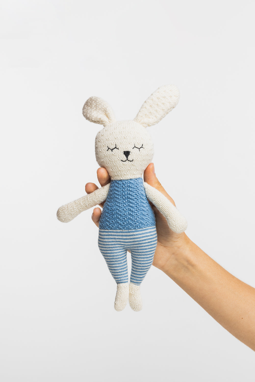 Hand holding a bunny soft toy wearing a blue outfit on a white background