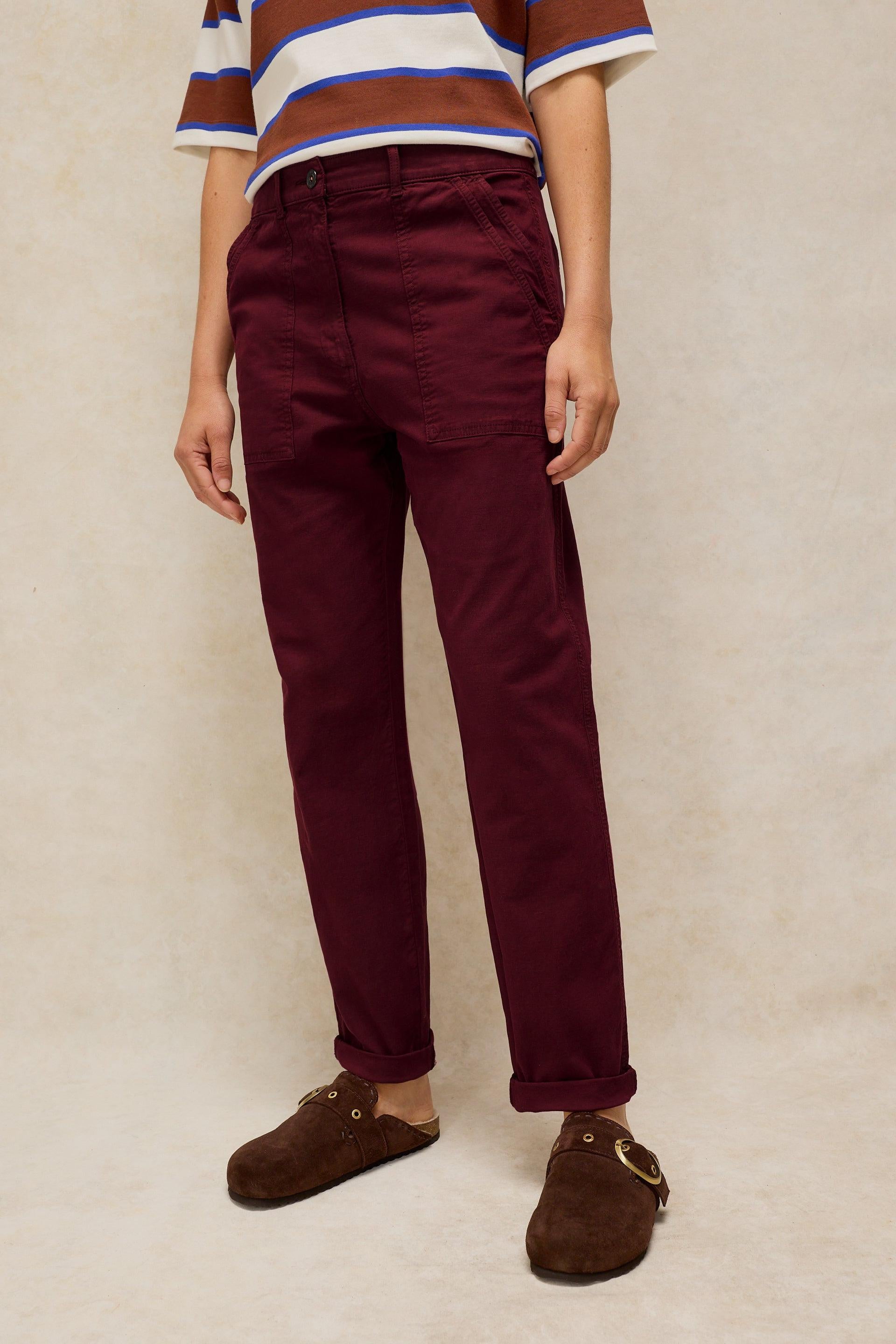 A person wearing burgundy organic cotton tapered trousers with a striped shirt and brown suede clogs.