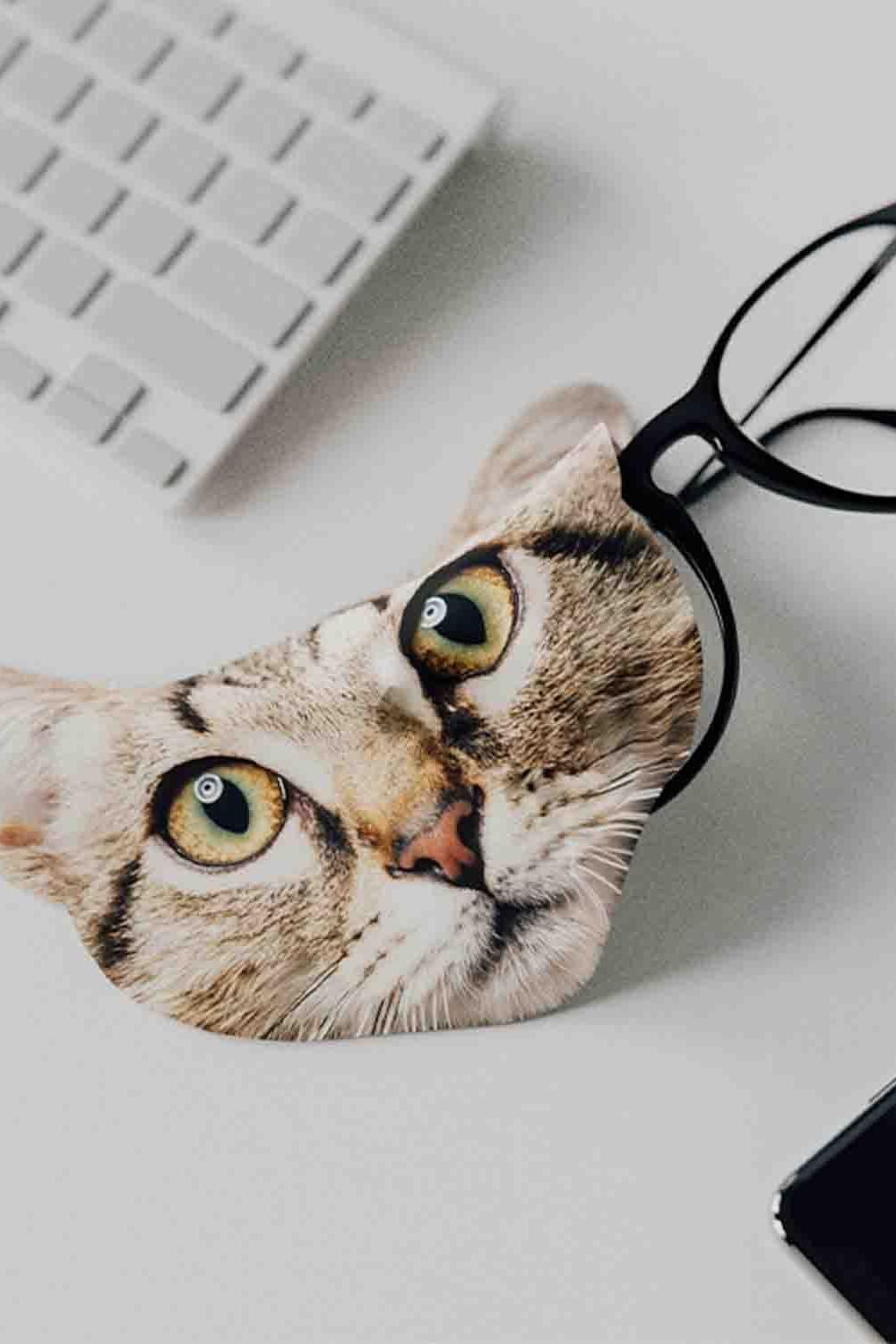 A close-up of a cat-themed eyeglass cloth with a pair of black-rimmed glasses on a white desk.