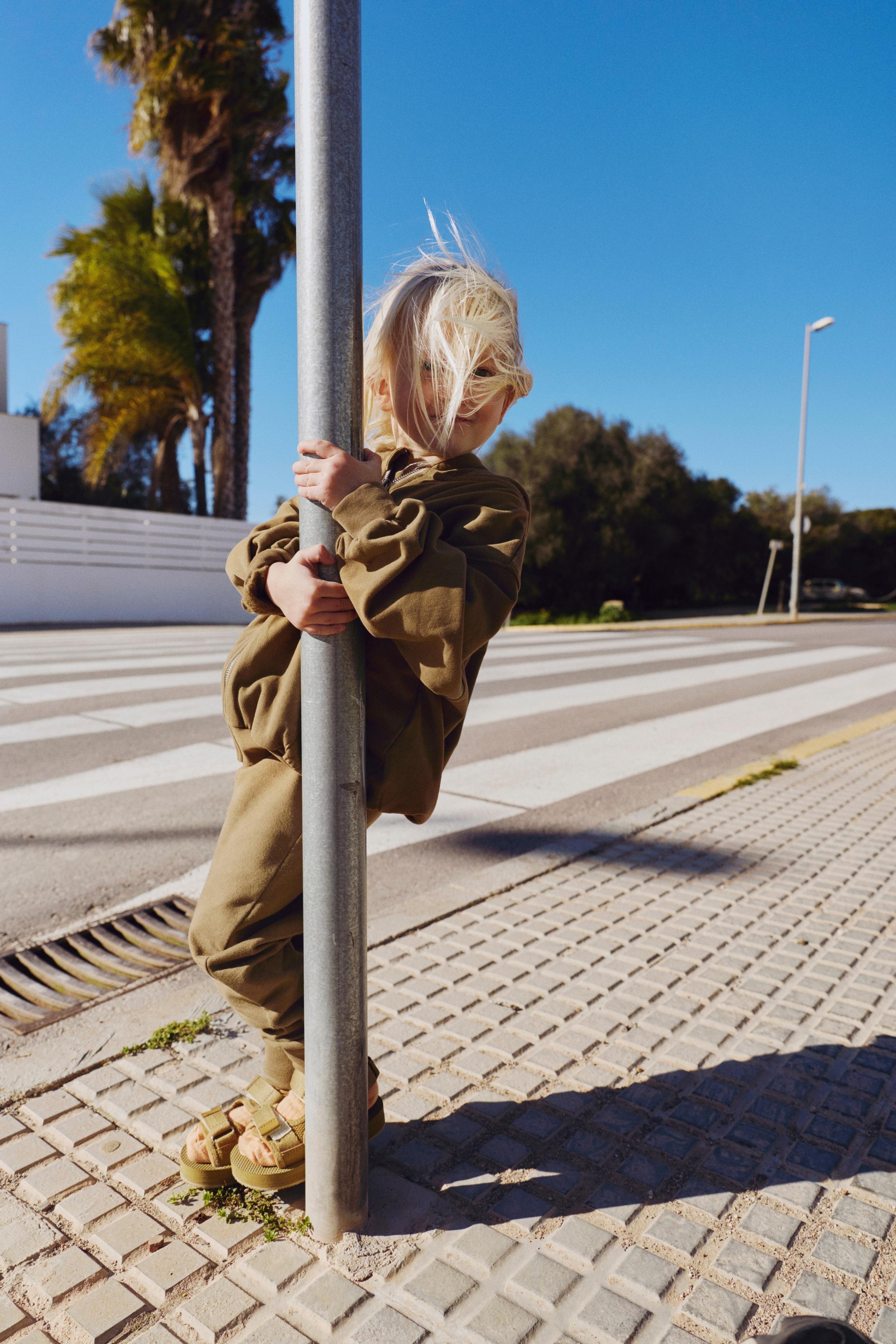 Child in a green outfit standing next to a pole on a sidewalk with trees and a building in the background.