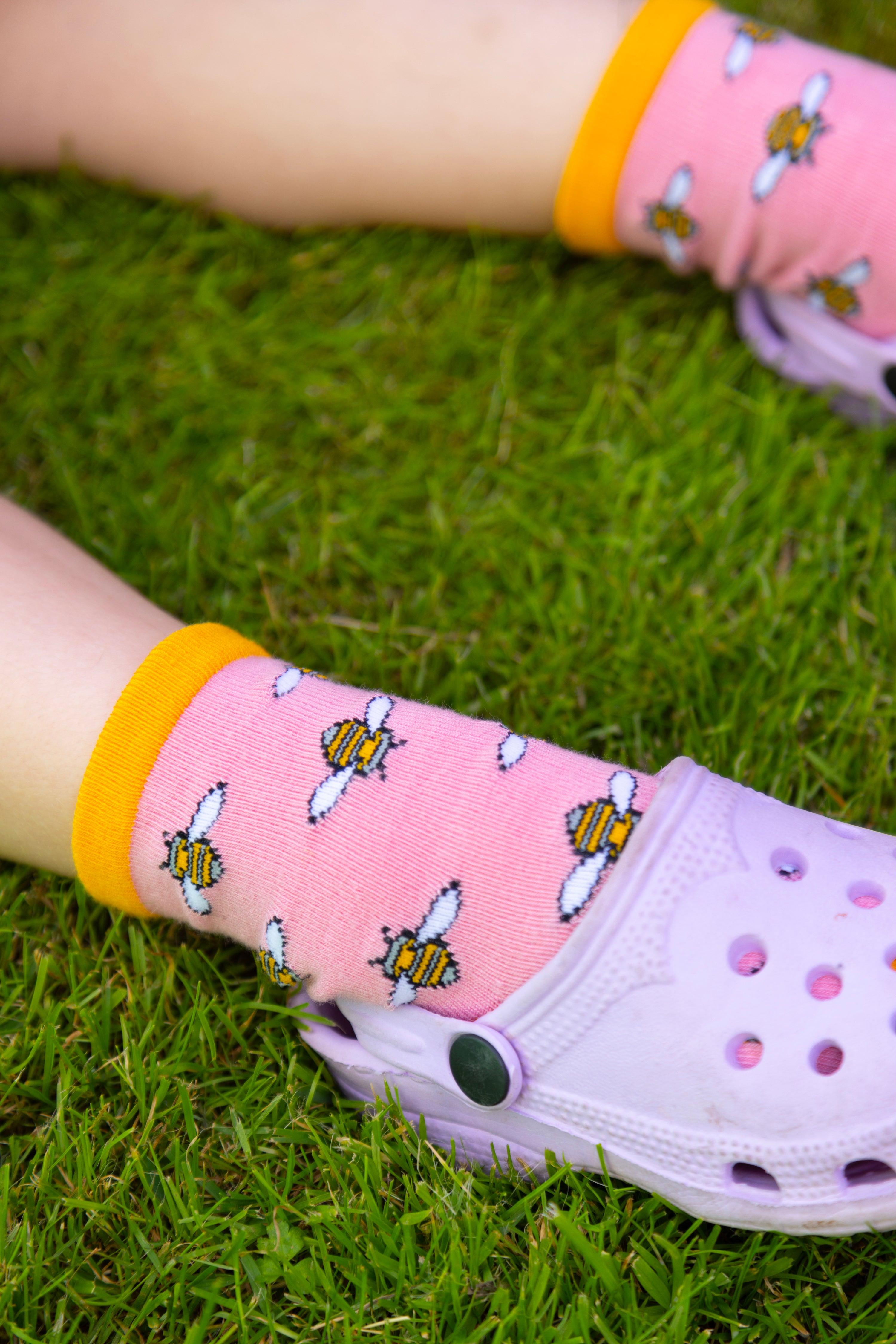 A child wearing pink bee-patterned socks and light purple clogs standing on green grass.