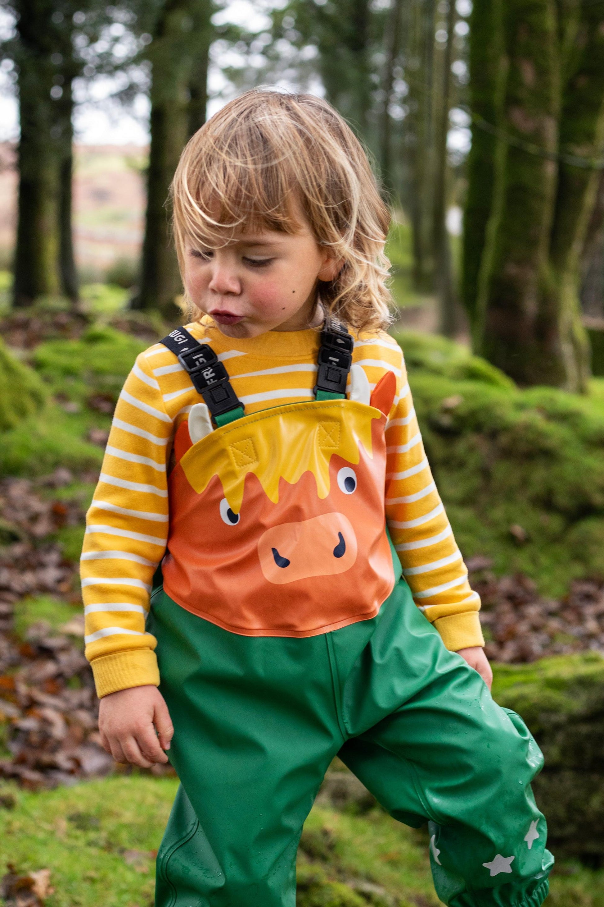 A toddler wearing Frugi highland cow dungarees and a yellow striped shirt in a mossy woodland setting