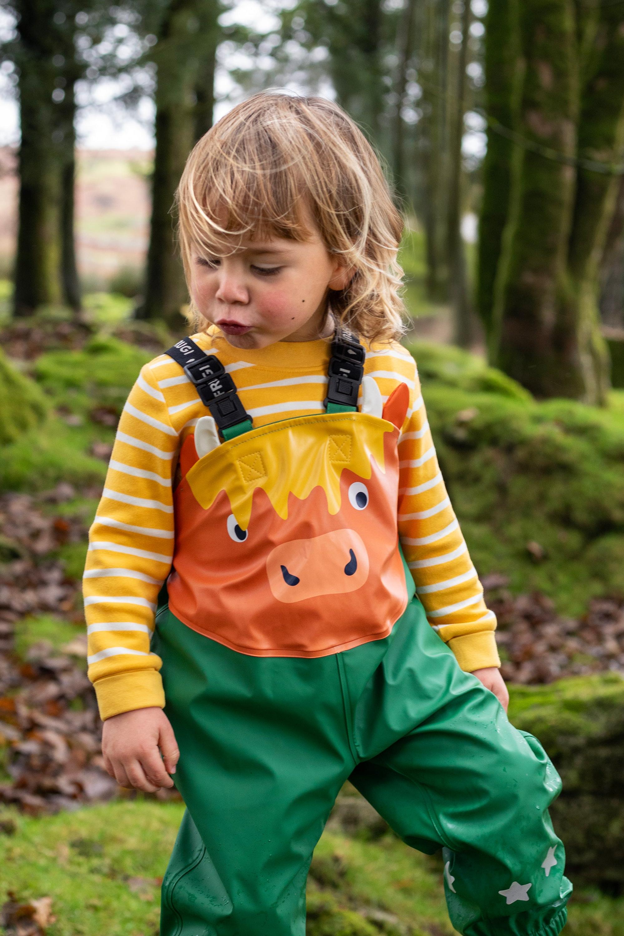 A toddler wearing Frugi highland cow dungarees and a yellow striped shirt in a mossy woodland setting