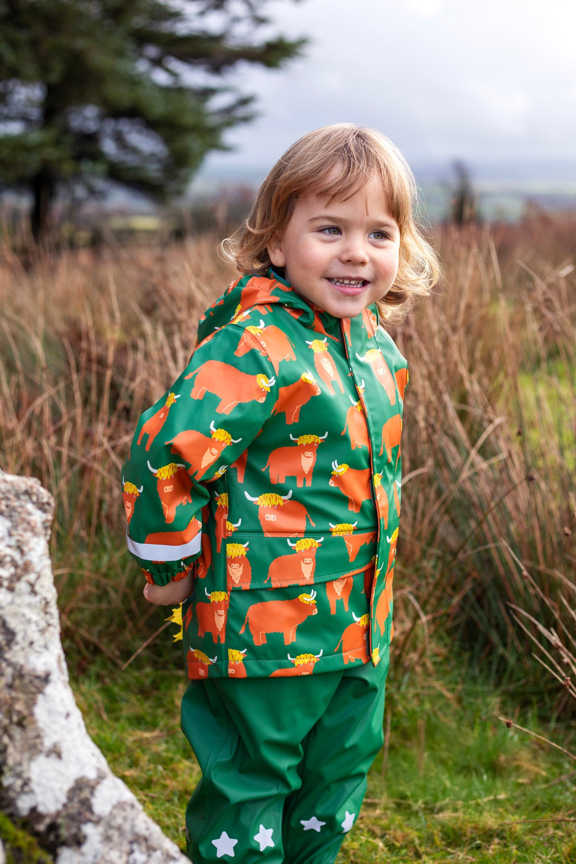A young child wearing a green Frugi waterproof raincoat with a highland cow print standing in a grassy field