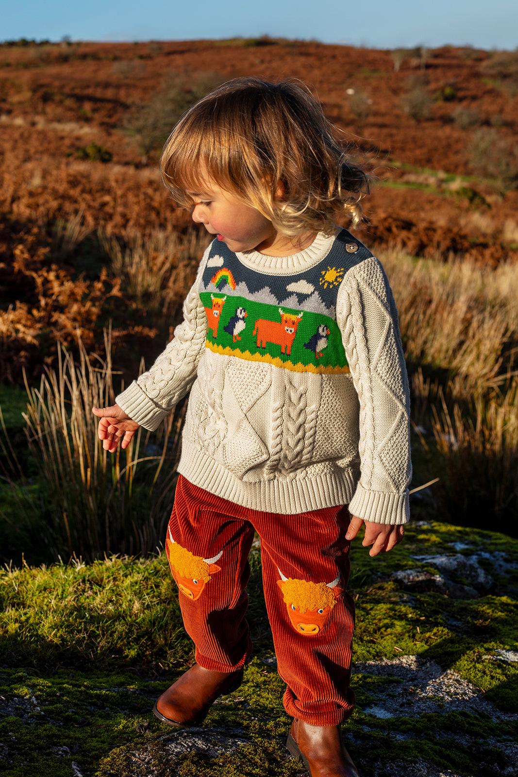 A child wearing a Frugi organic cotton knit jumper with animal patterns, standing in a grassy field during sunset.