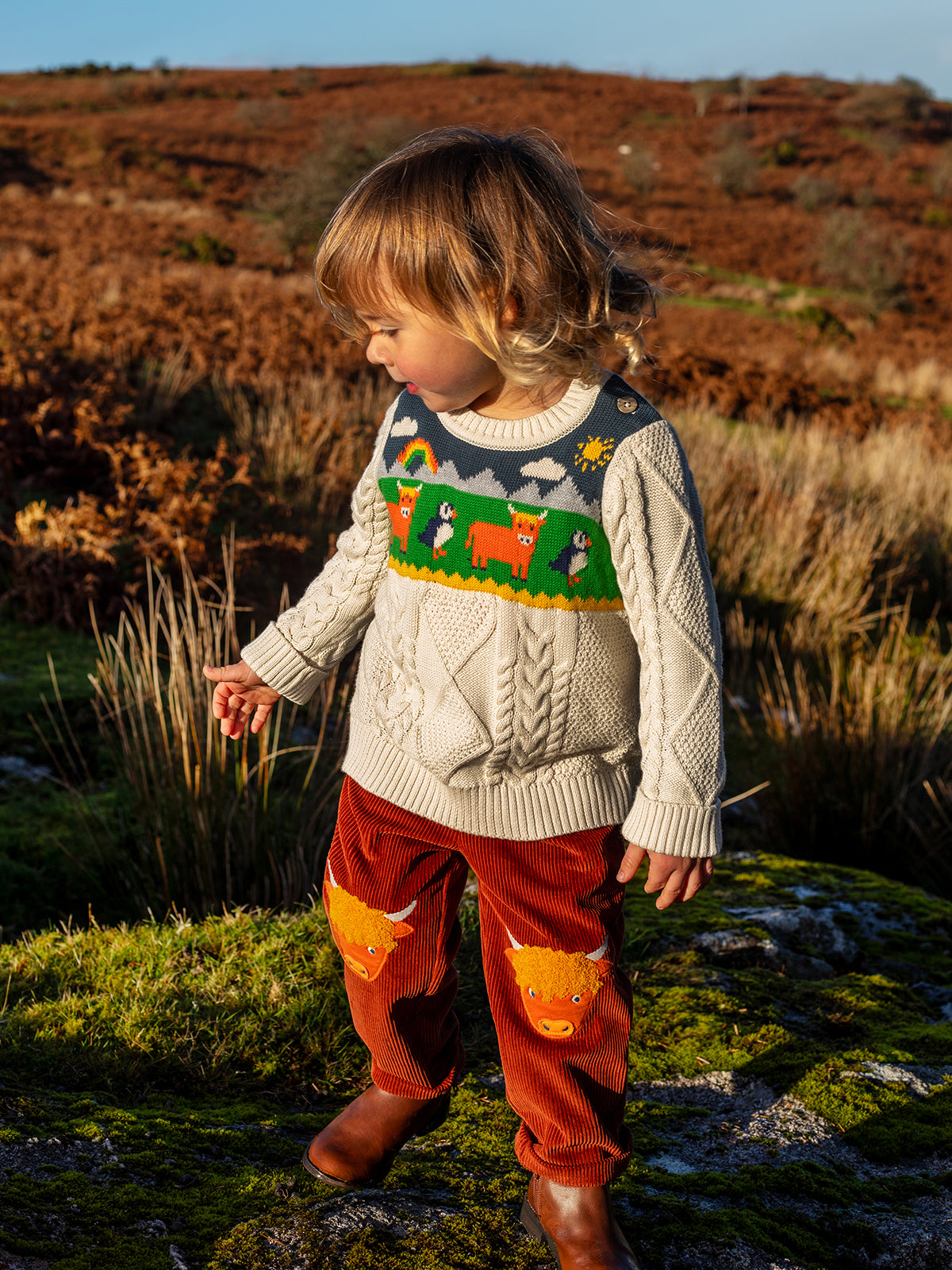 A child wearing a Frugi organic cotton knit jumper with animal patterns, standing in a grassy field during sunset.