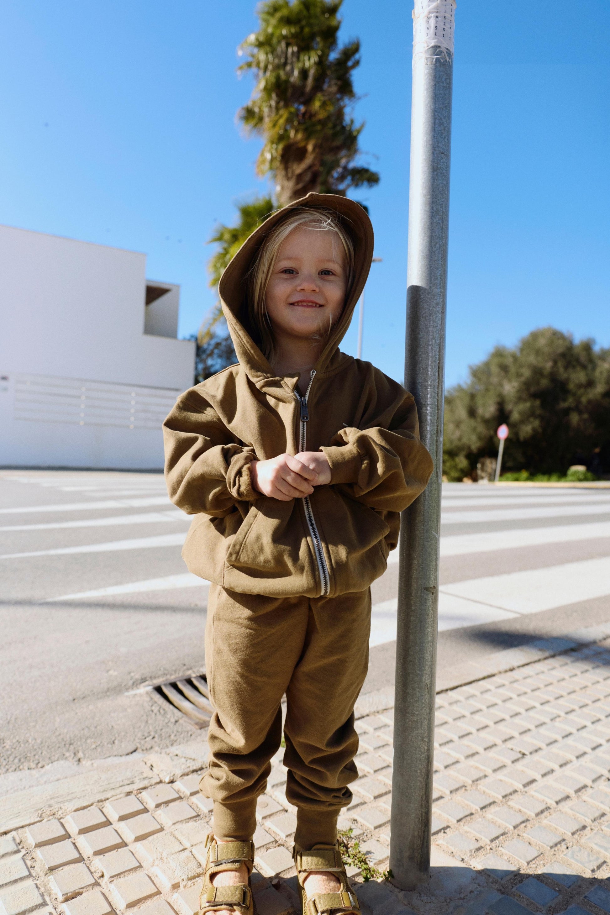 Child wearing a brown hoodie and pants standing on a sidewalk with trees and buildings in the background.