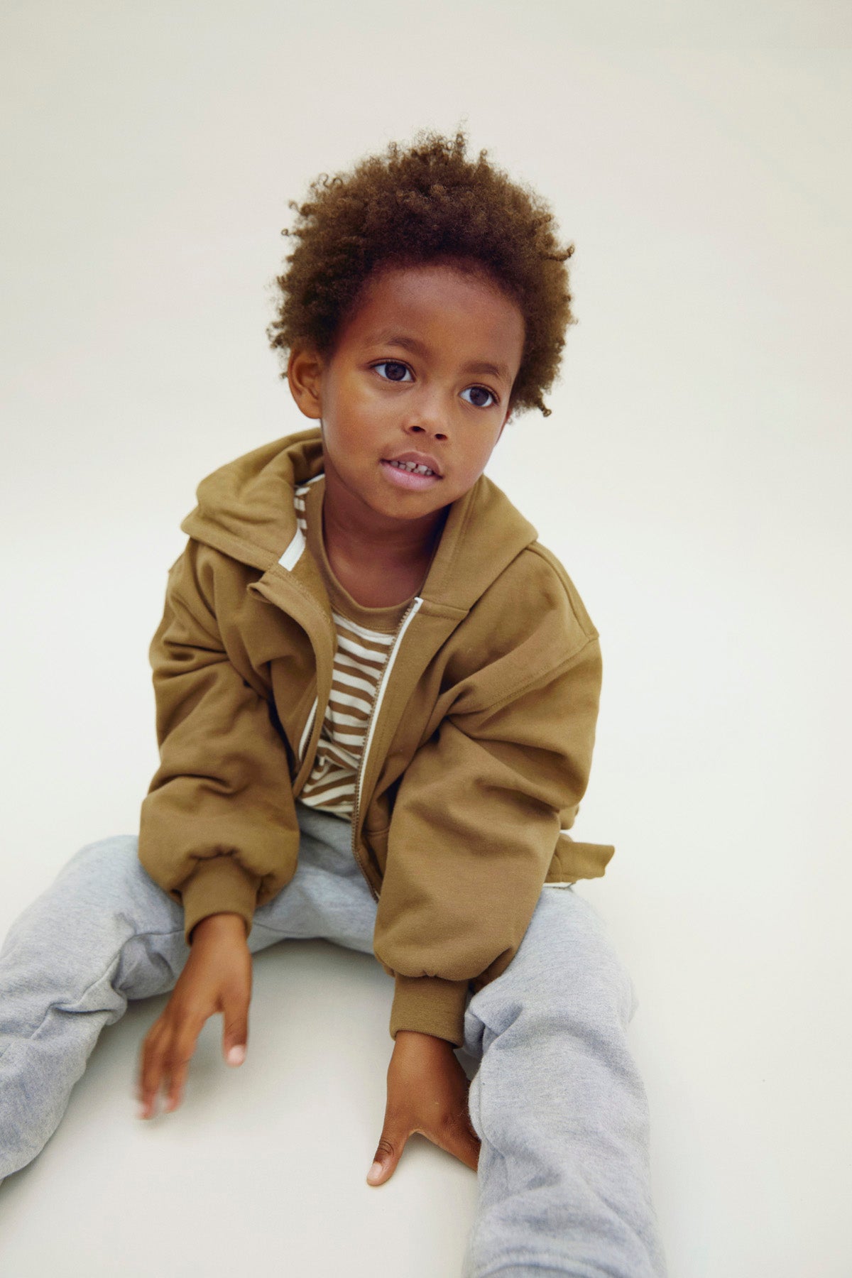 Child wearing a brown organic cotton hoodie and gray pants sitting on a white surface.