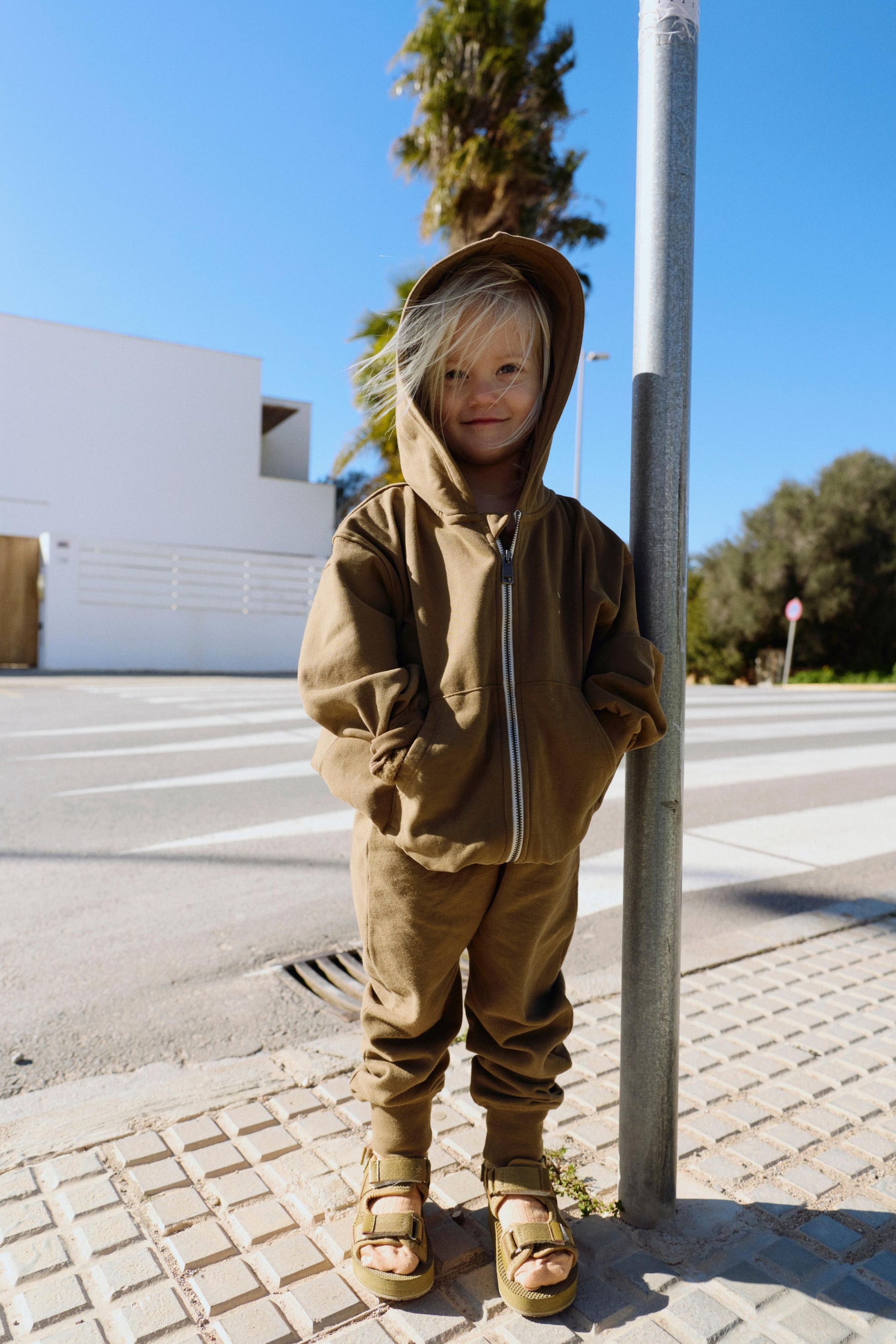 Child in a brown hooded outfit standing on a sidewalk with a streetlight and palm tree in the background.