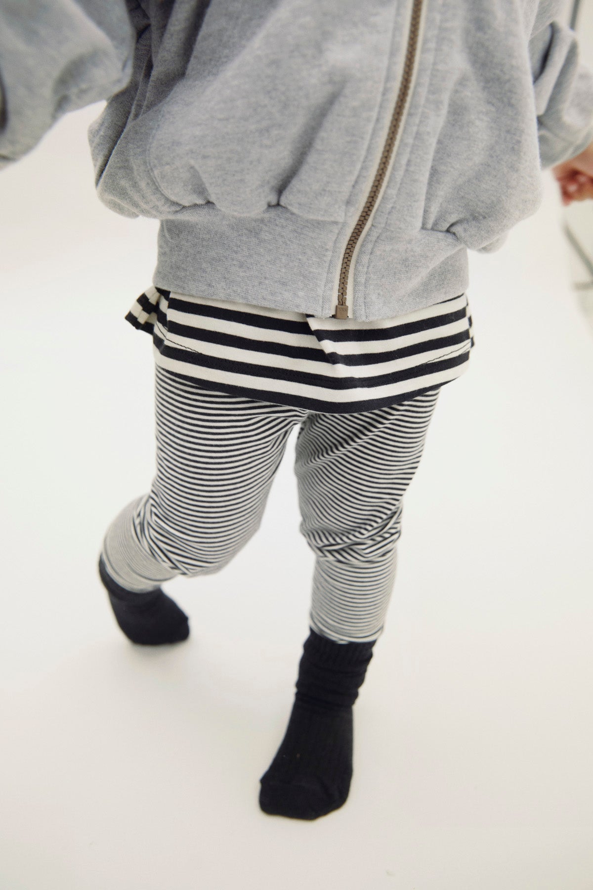 A child wearing a light gray zip-up hoodie, black and white striped t-shirt, and black and white striped pants with black socks, shown from behind against a white background.