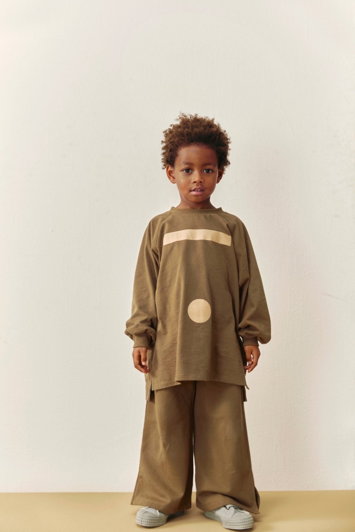 A young child stands against a plain off-white wall wearing a loose, oversized two-piece outfit in a muted taupe-brown color with a horizontal beige stripe across the chest and a beige circle detail on the front. The outfit includes wide-leg pants and light gray sneakers.