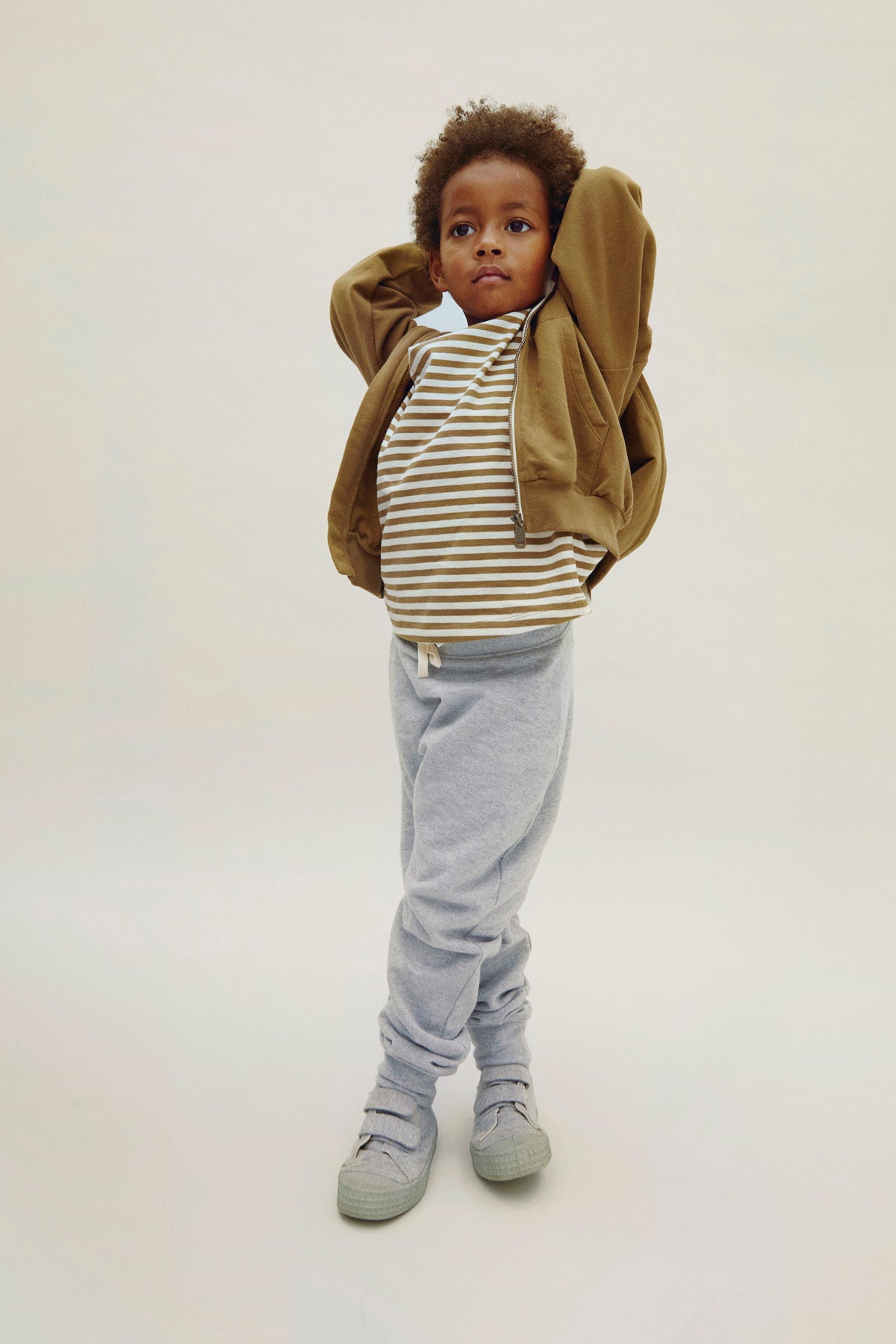 Child wearing a brown hoodie, striped shirt, and gray pants on a white background