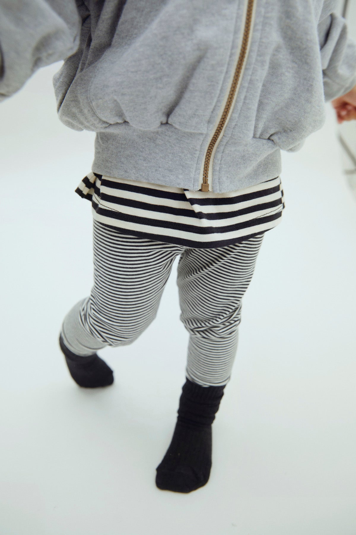 A child wearing a light gray zip-up hoodie, black and white striped t-shirt, and black and white striped pants with black socks, shown from behind against a white background.