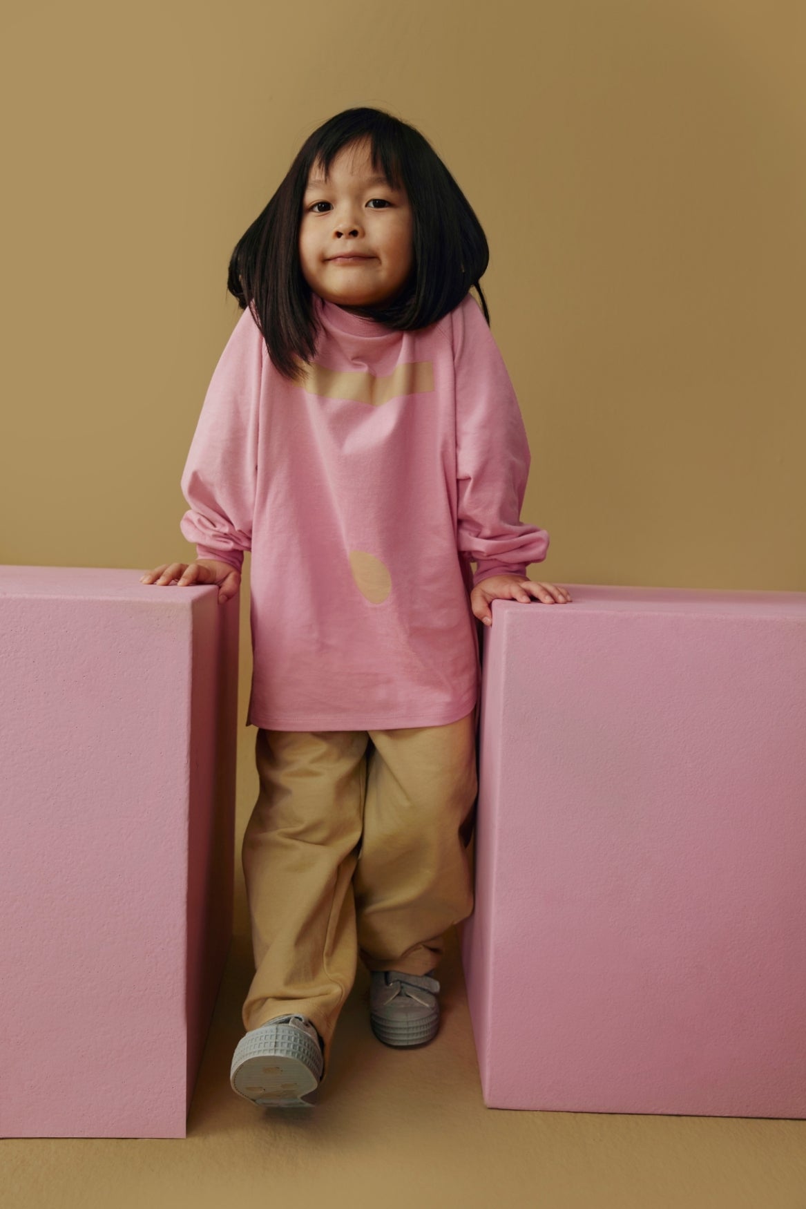A young child with short dark hair stands between two large pink blocks against a warm tan background, wearing an oversized pink long-sleeve top with a small yellow graphic, tan pants, and gray sneakers.