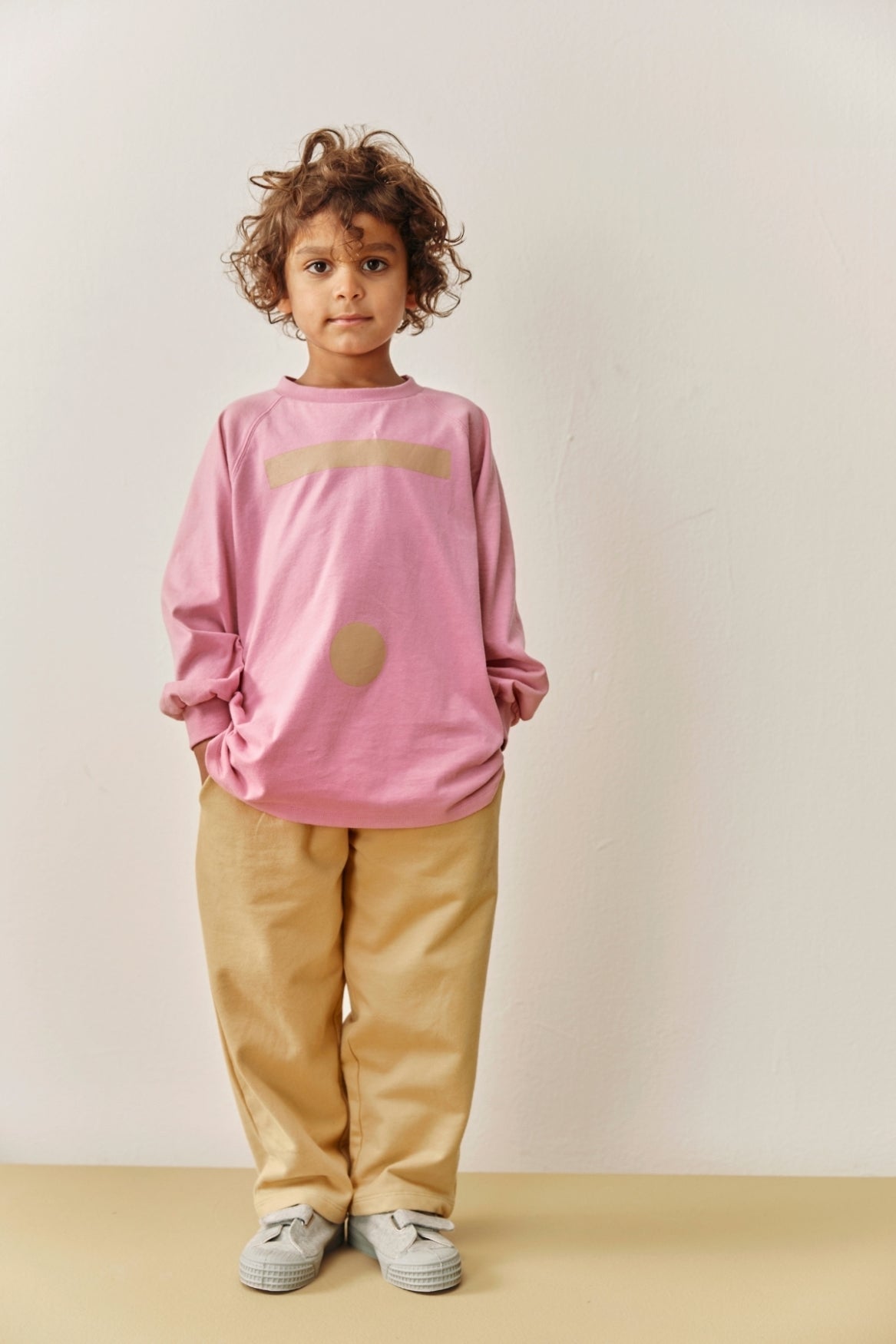 A young child with curly brown hair stands against a plain off-white wall wearing a pink long-sleeve t-shirt with a beige graphic, beige pants, and light gray sneakers.