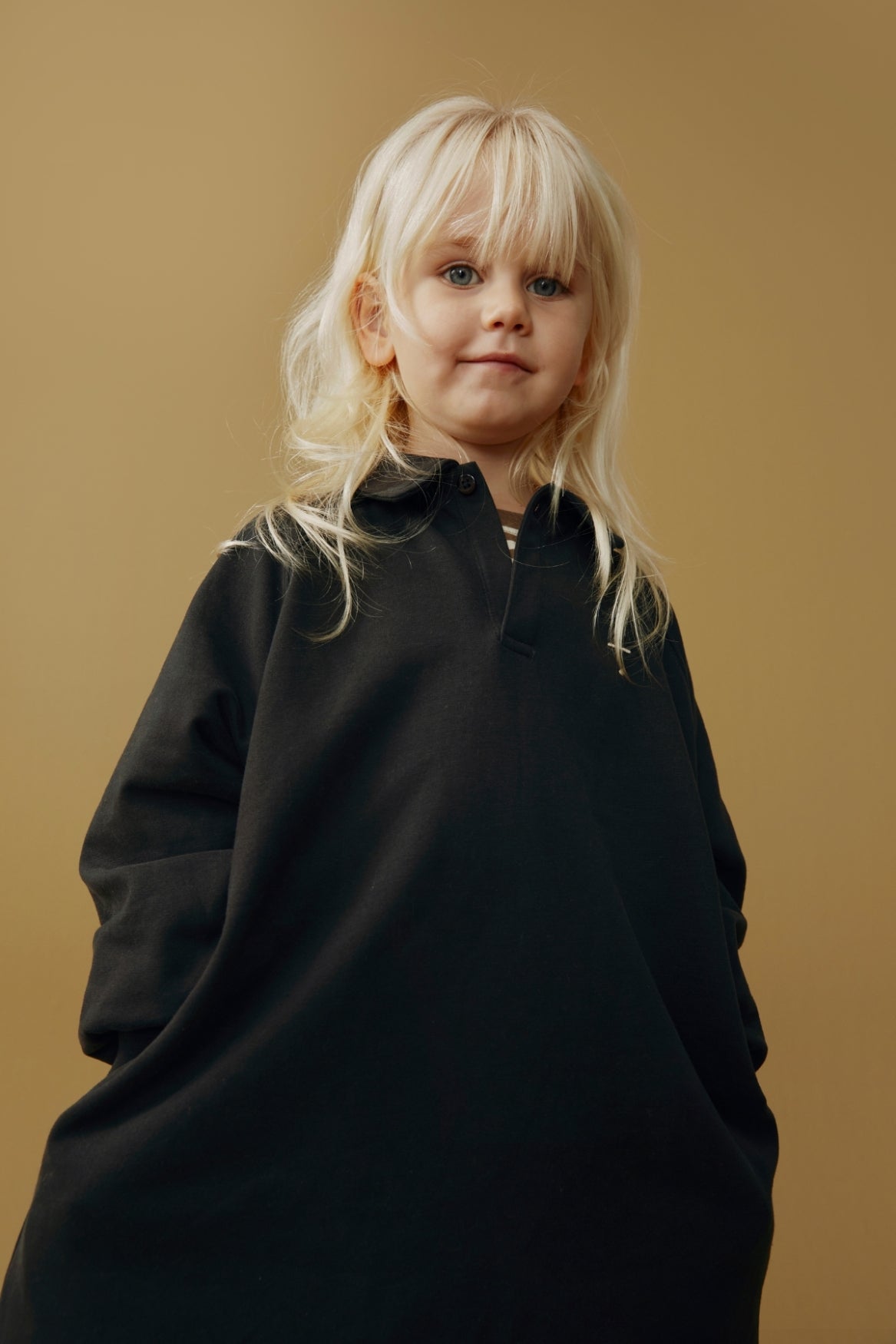 A young child with blonde hair wearing an oversized black long-sleeve dress, photographed against a solid warm tan background.