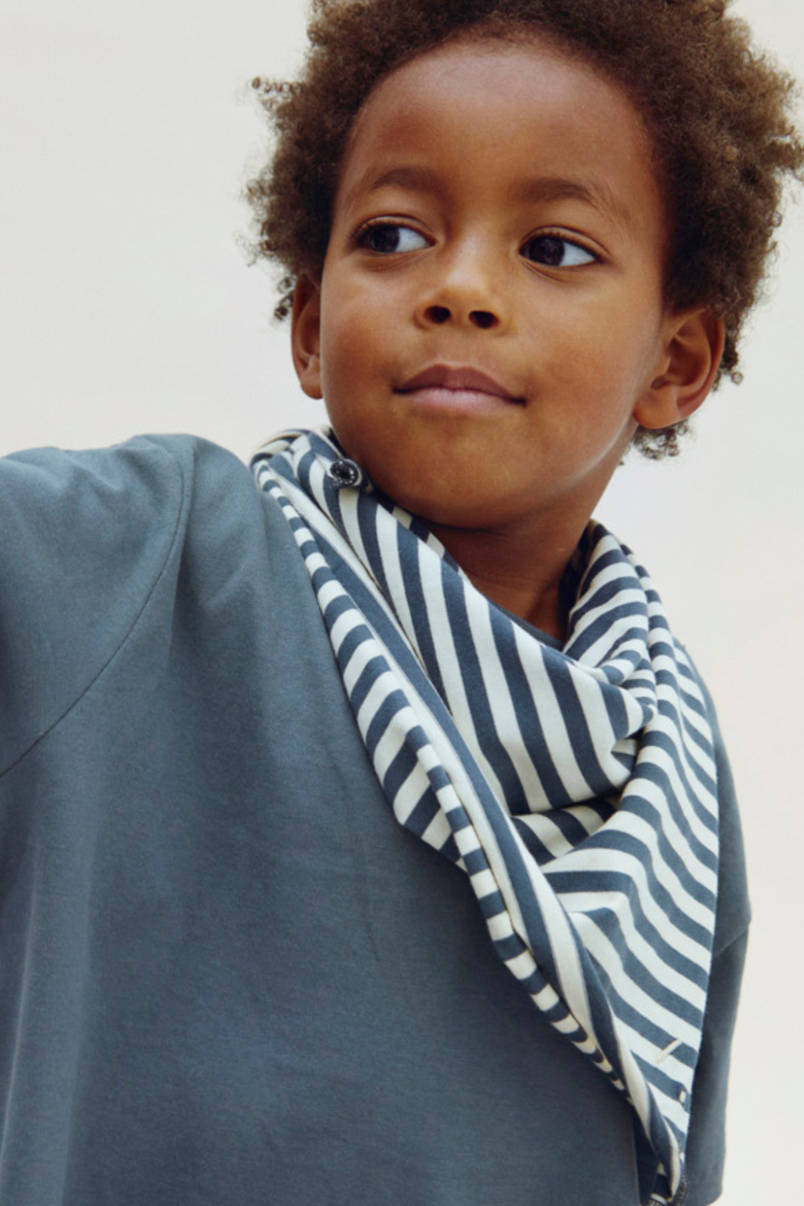A close-up of a young child wearing a navy and white striped scarf and a slate blue-gray top against a plain light background.