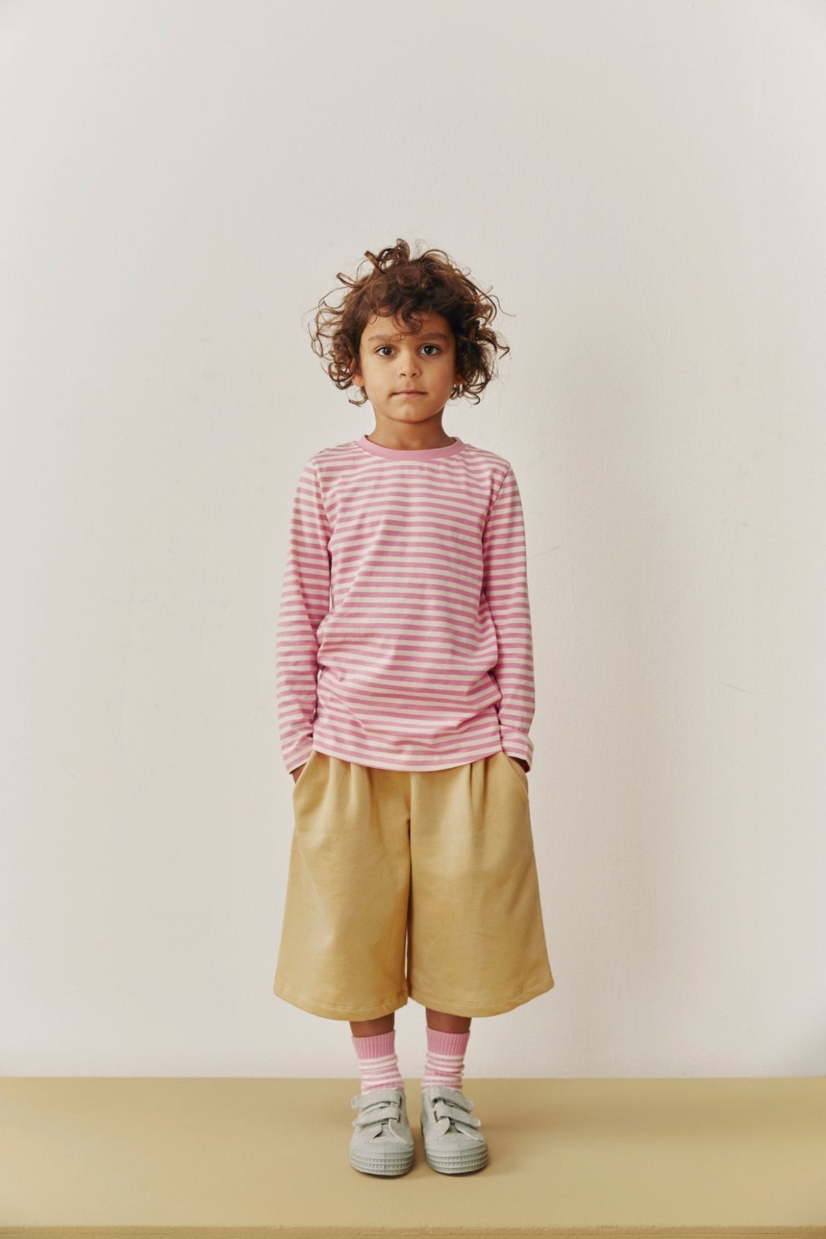 Child wearing a pink striped shirt and beige shorts against a white background