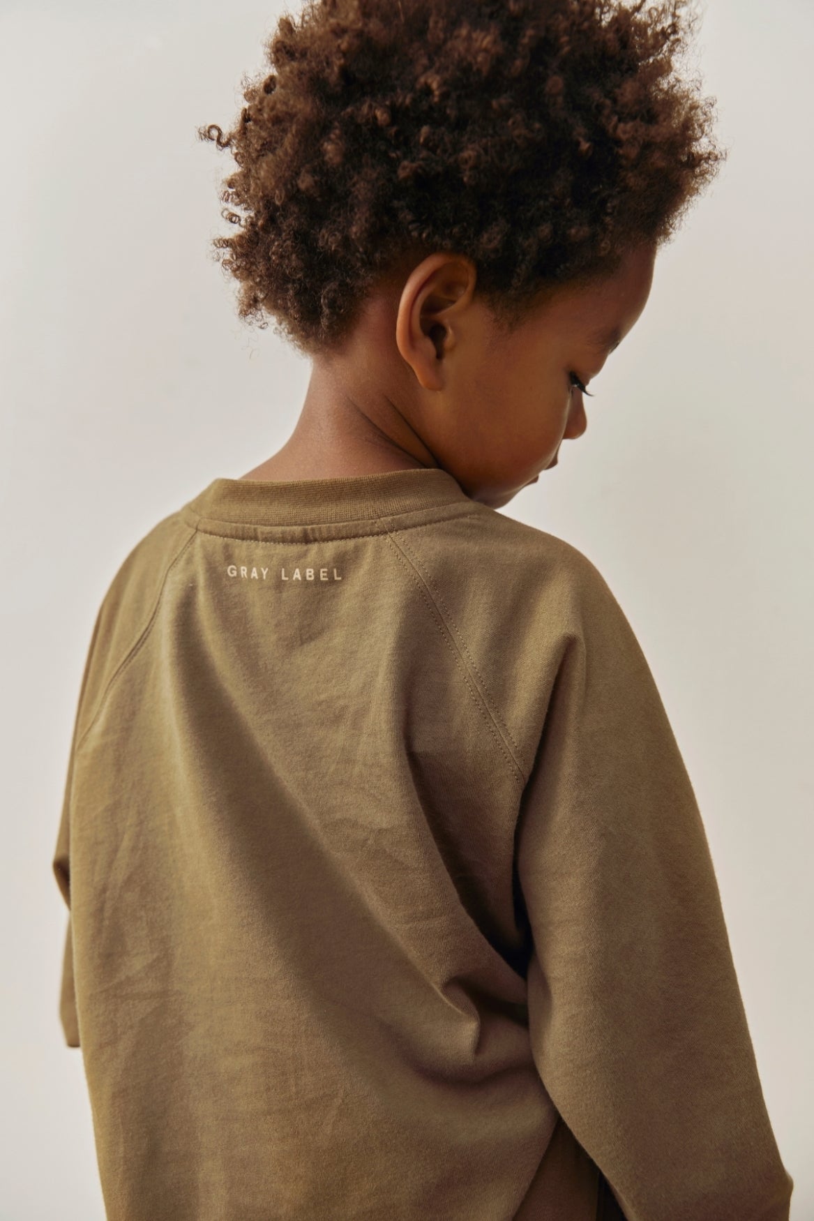 A child with curly brown hair seen from the back, wearing a muted brownish-green long-sleeve sweatshirt with 'GRAY LABEL' printed in small letters on the upper back, against a plain light background.