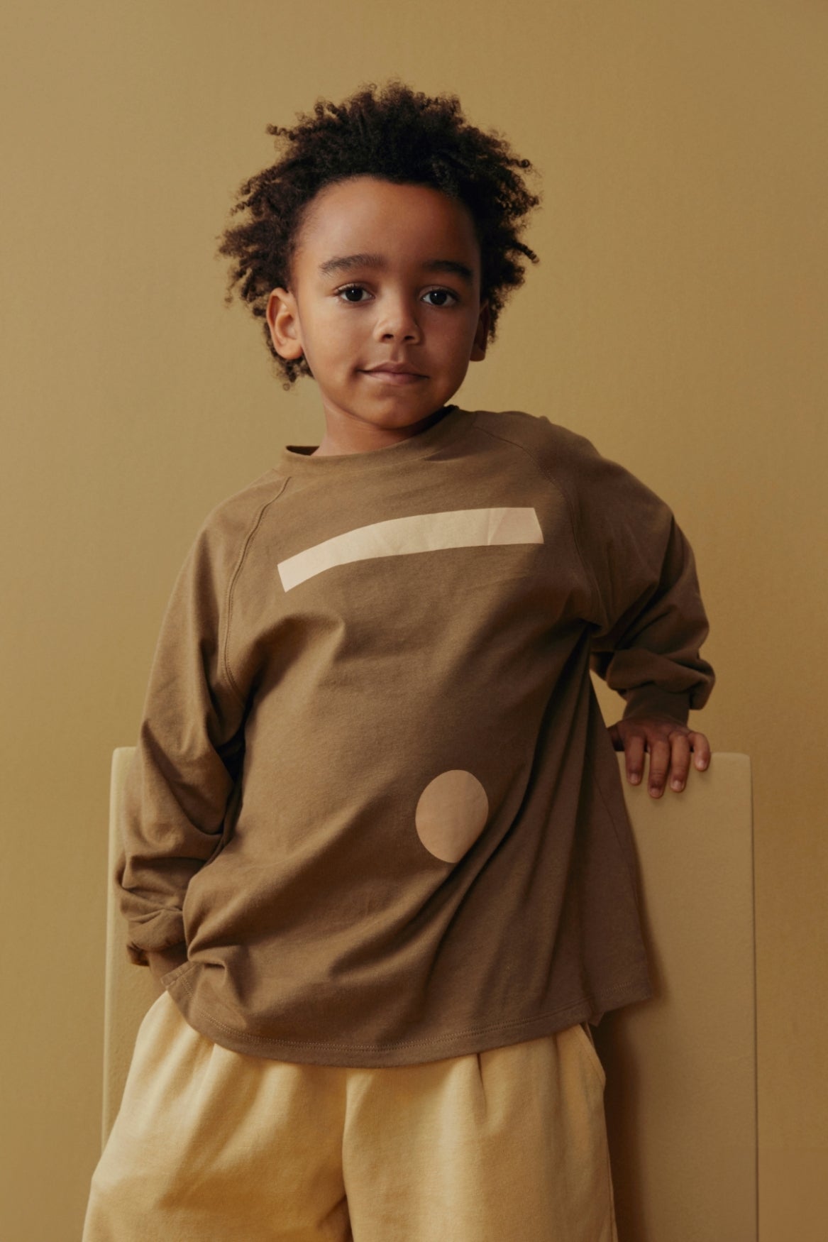 A young child with curly dark hair wearing a brown long-sleeve top with a cream stripe and circle graphic, paired with light beige pants, standing against a solid beige background.