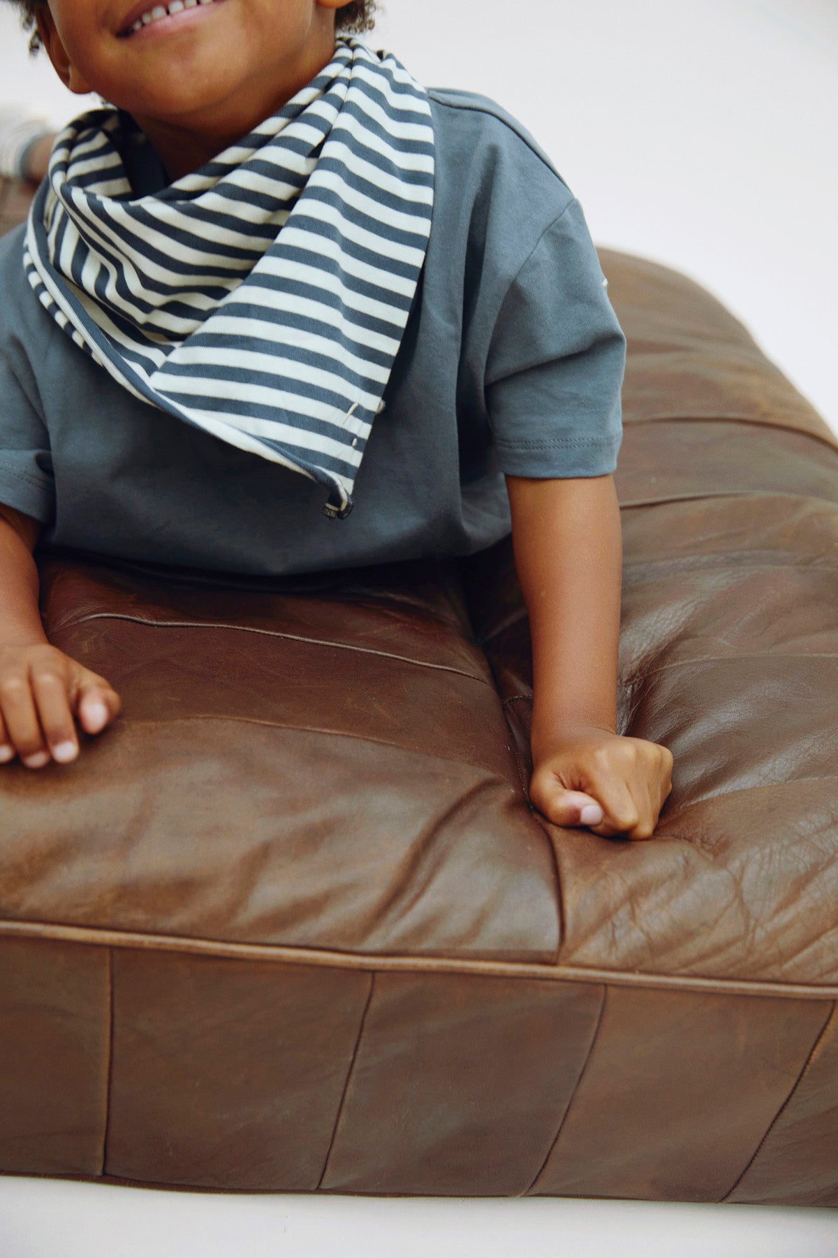 A child wearing a slate blue-gray t-shirt and a navy and white striped bandana bib, sitting on a brown leather cushion.