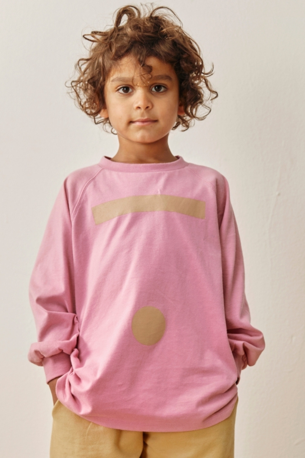A young child with curly brown hair wearing a pink long-sleeve sweatshirt with a beige arched stripe and a beige oval patch on the front, paired with tan pants, against a plain off-white wall.