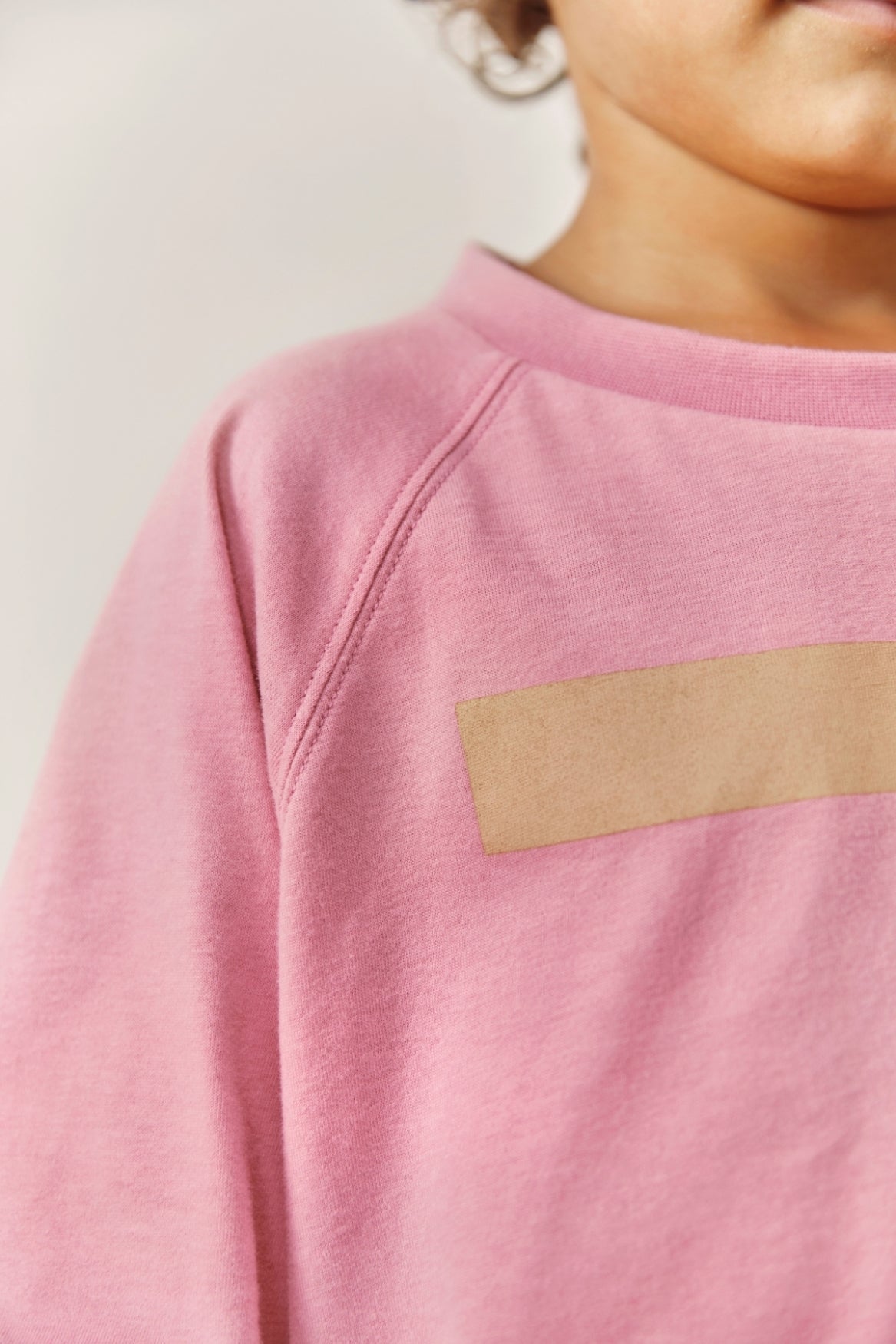 Close-up of a child wearing a pink sweatshirt with a horizontal beige patch detail across the chest.