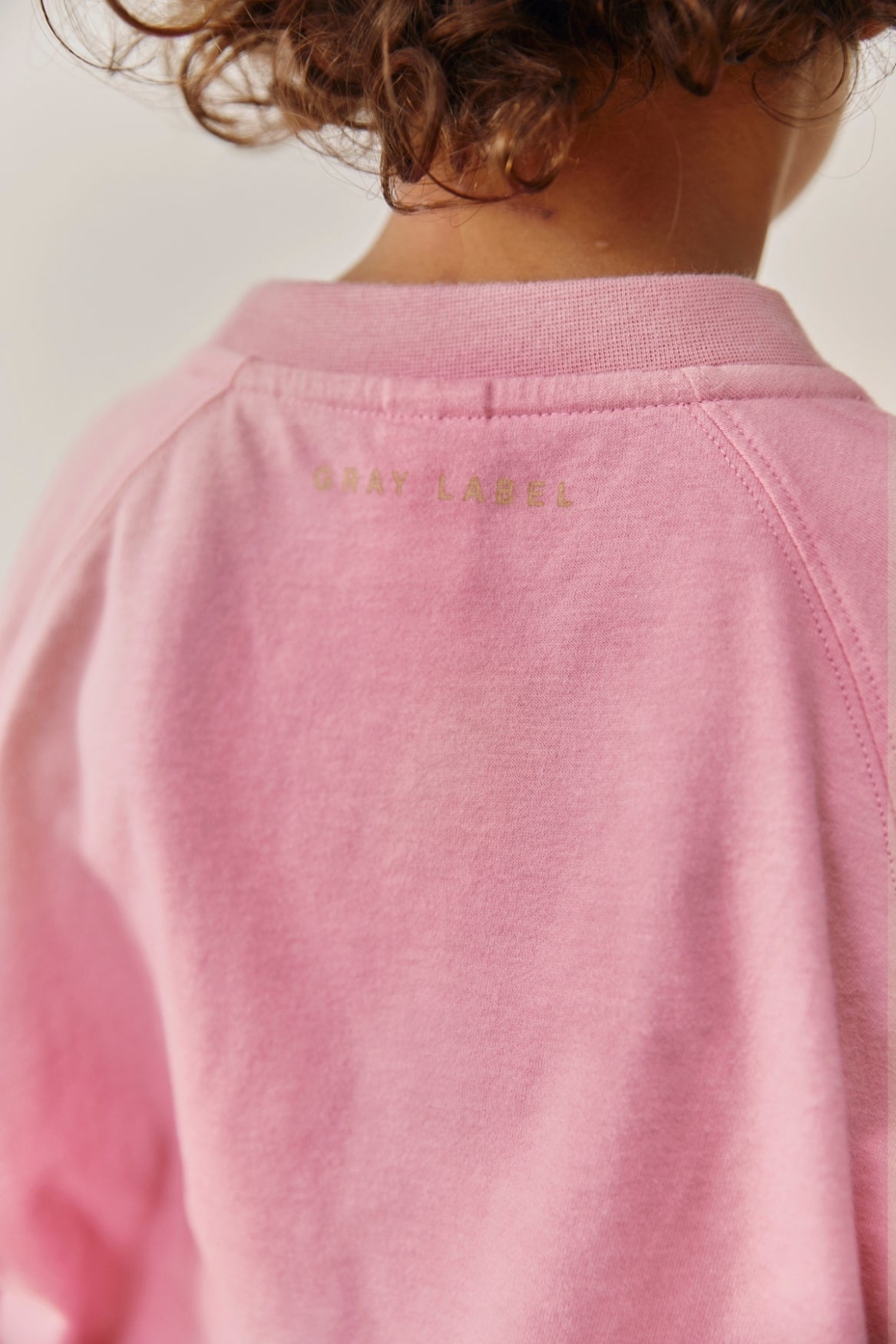 Close-up back view of a child wearing a light pink sweatshirt, showing the inner collar with the brand name 'GRAY LABEL' printed in gold.