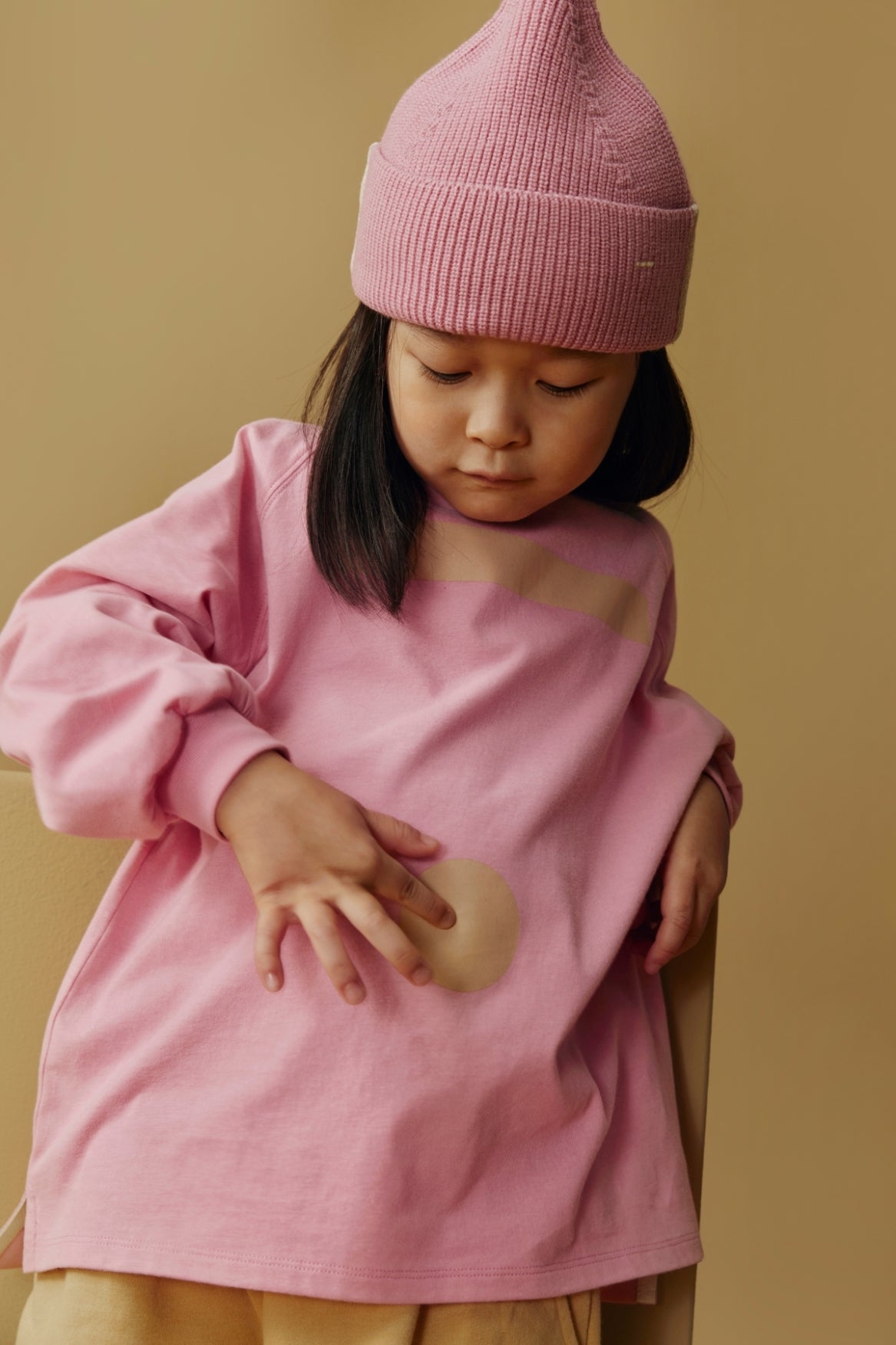 A young child wearing a pink knit beanie and a pink oversized long-sleeve top, posed against a warm beige background.