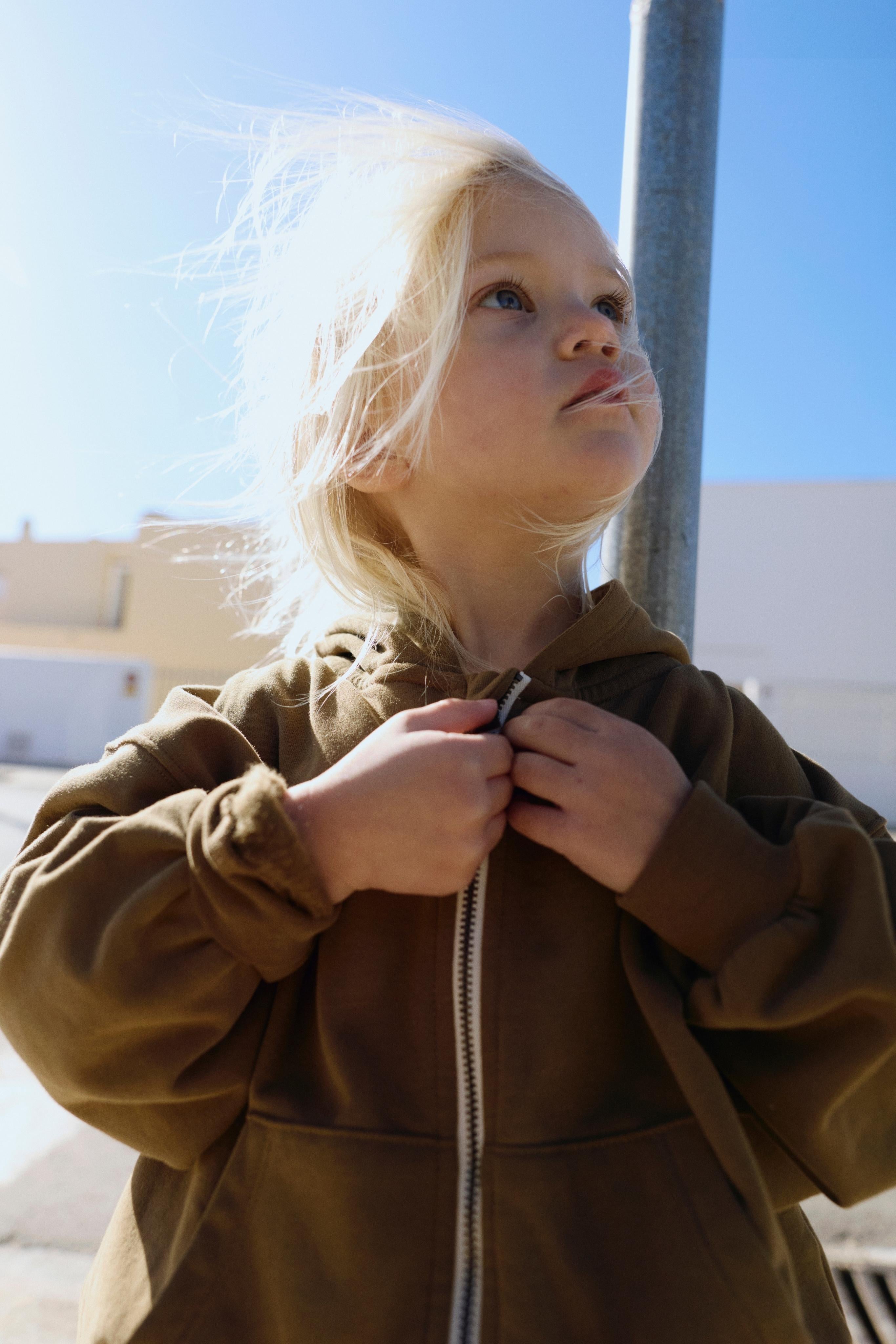 Child wearing a brown coat with a hood, standing outdoors on a sunny day.