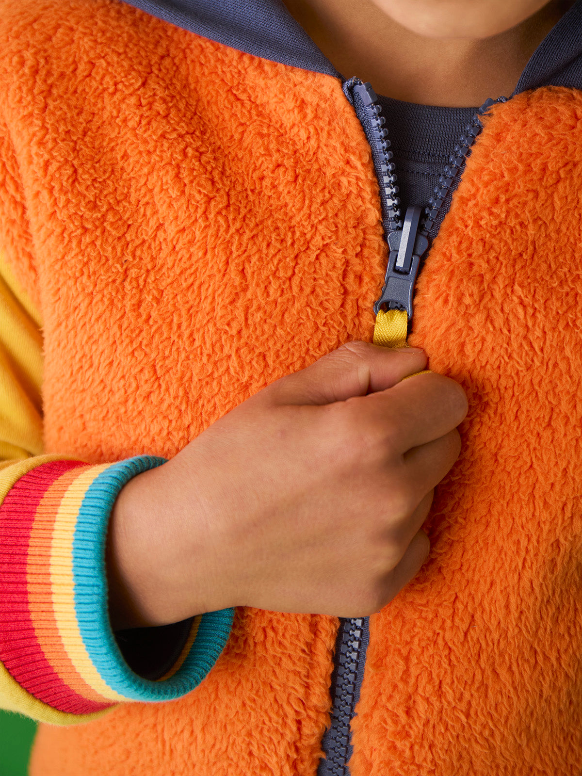 Child zipping up a bright orange Frugi organic cotton hoody with colorful striped cuffs.