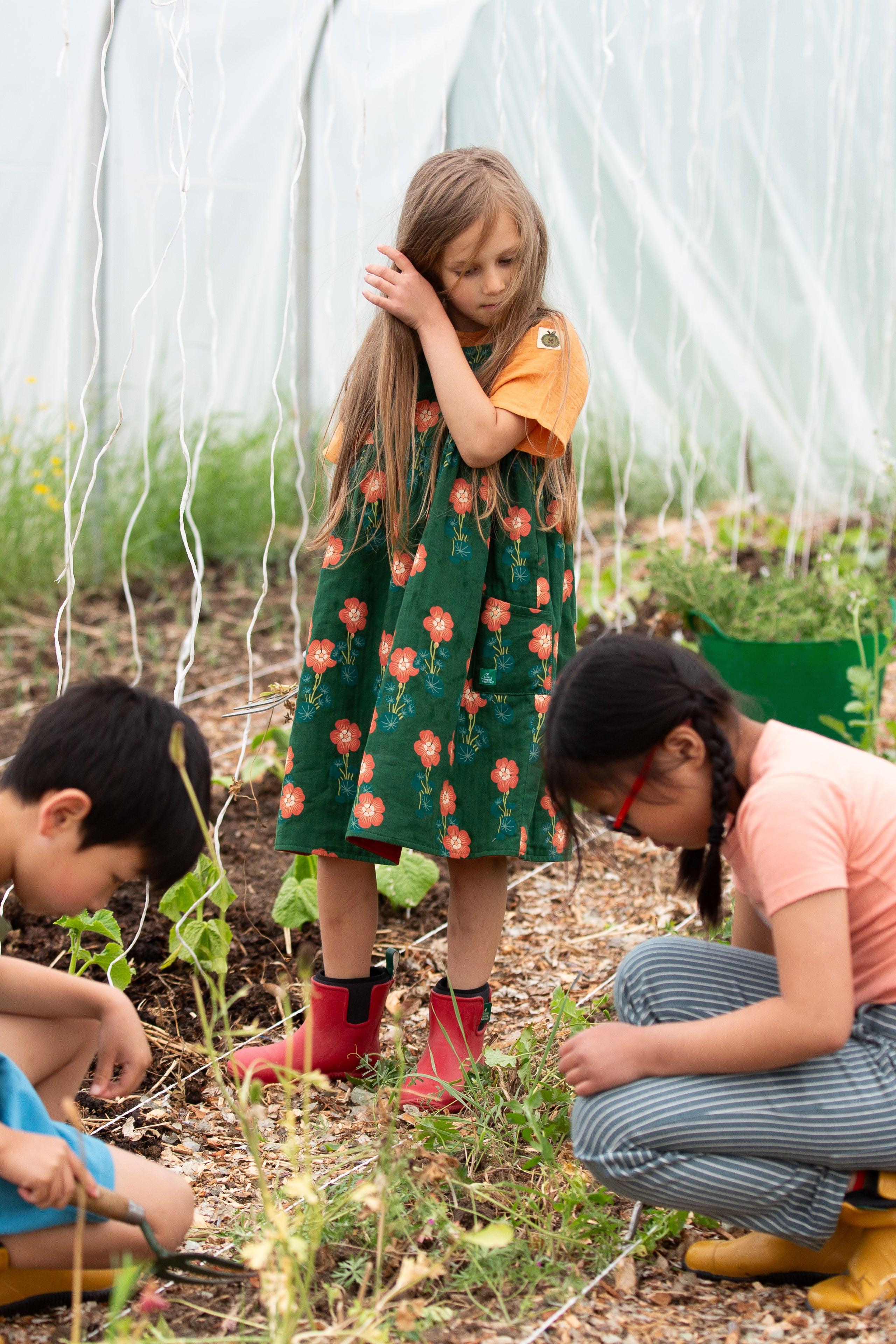 Children in a garden, with one child in a green dress and red boots standing and two others in yellow rain boots crouching.