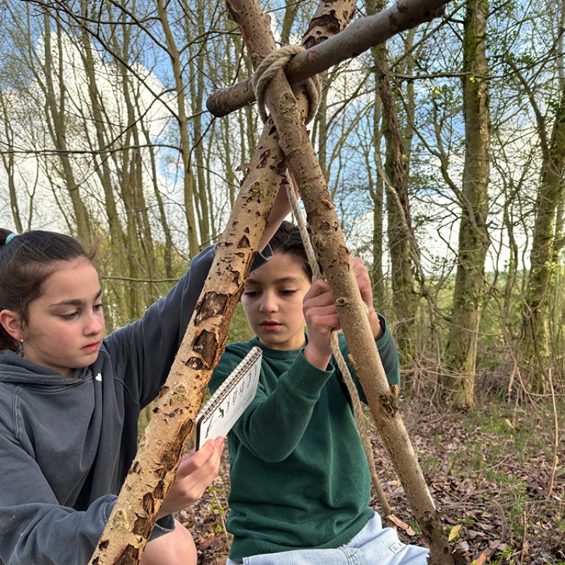 Two children outside in a forest learning to tie ropes between sticks using an instruction manual.