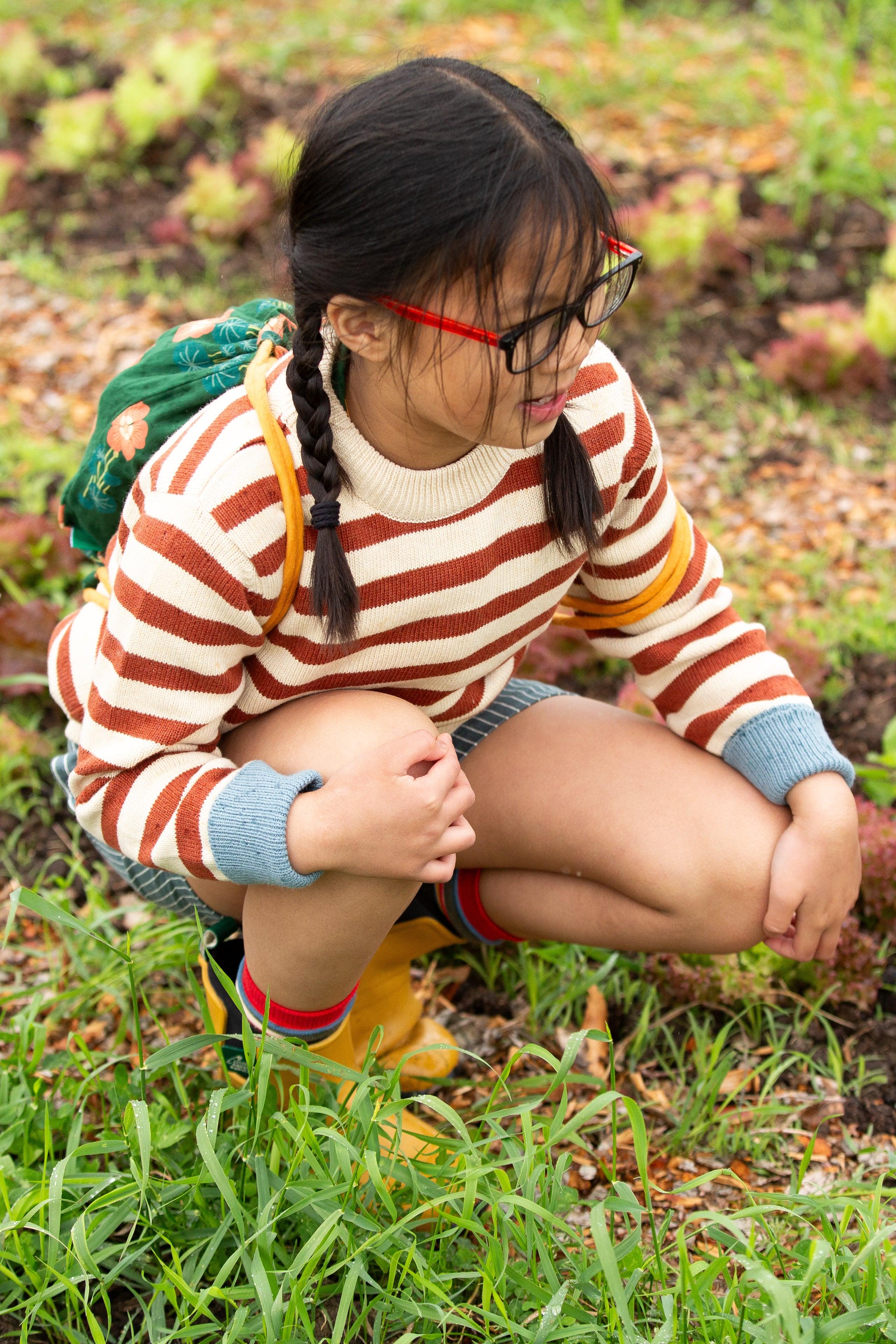 Young girl in a striped Little Green Radicals knit sweater and rain boots crouching in a garden
