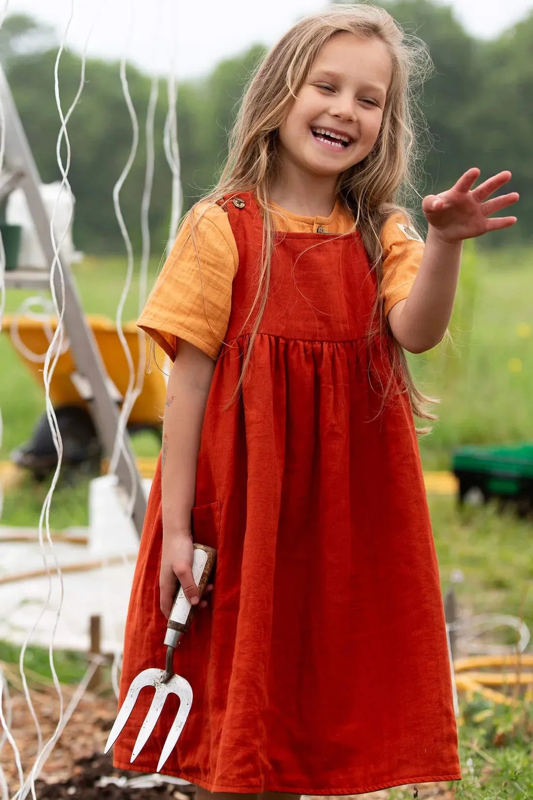 Young girl in a red organic cotton dress and yellow shirt holding a garden fork, standing in a field.