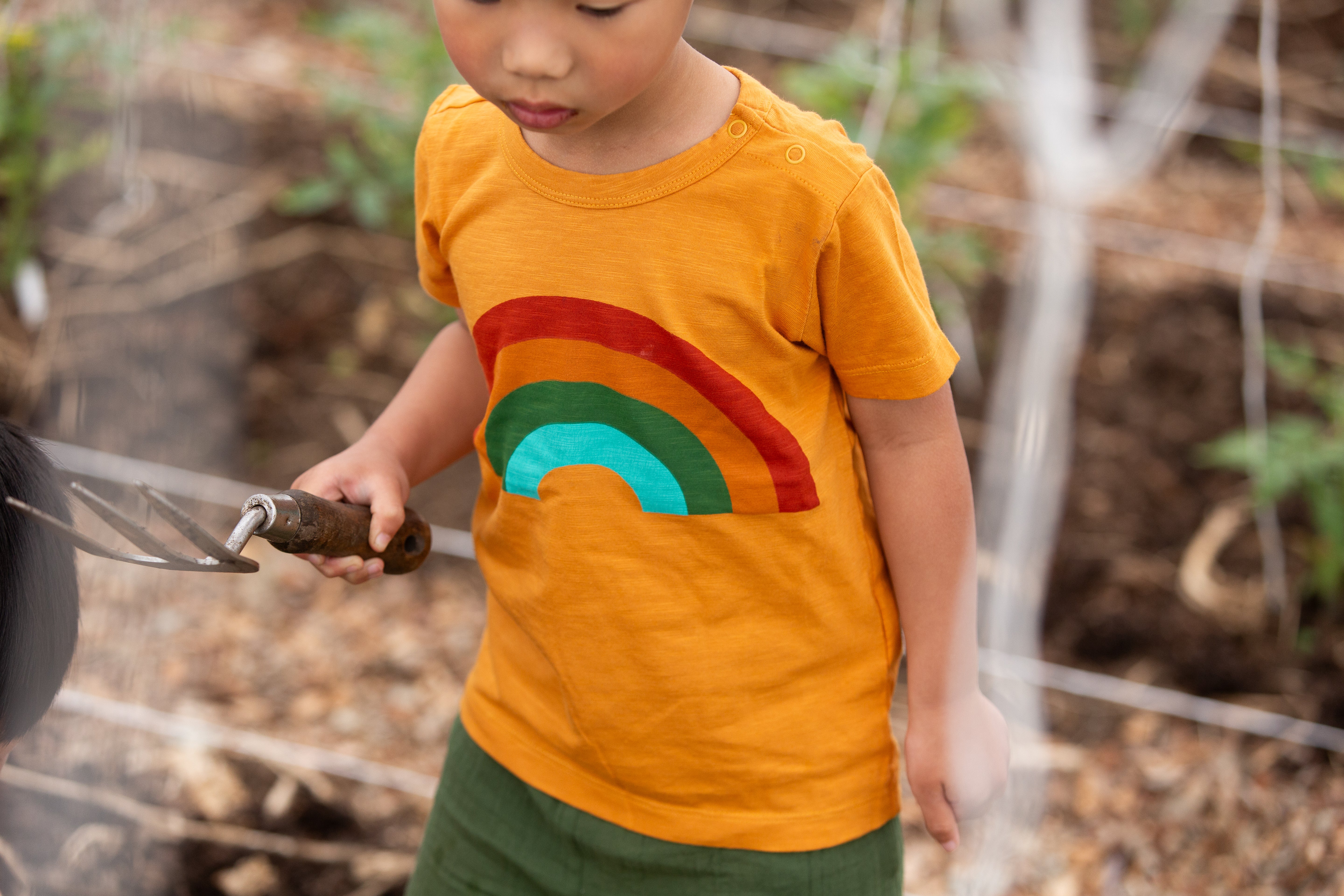 Child wearing an orange shirt with a rainbow design, holding a tool outdoors.