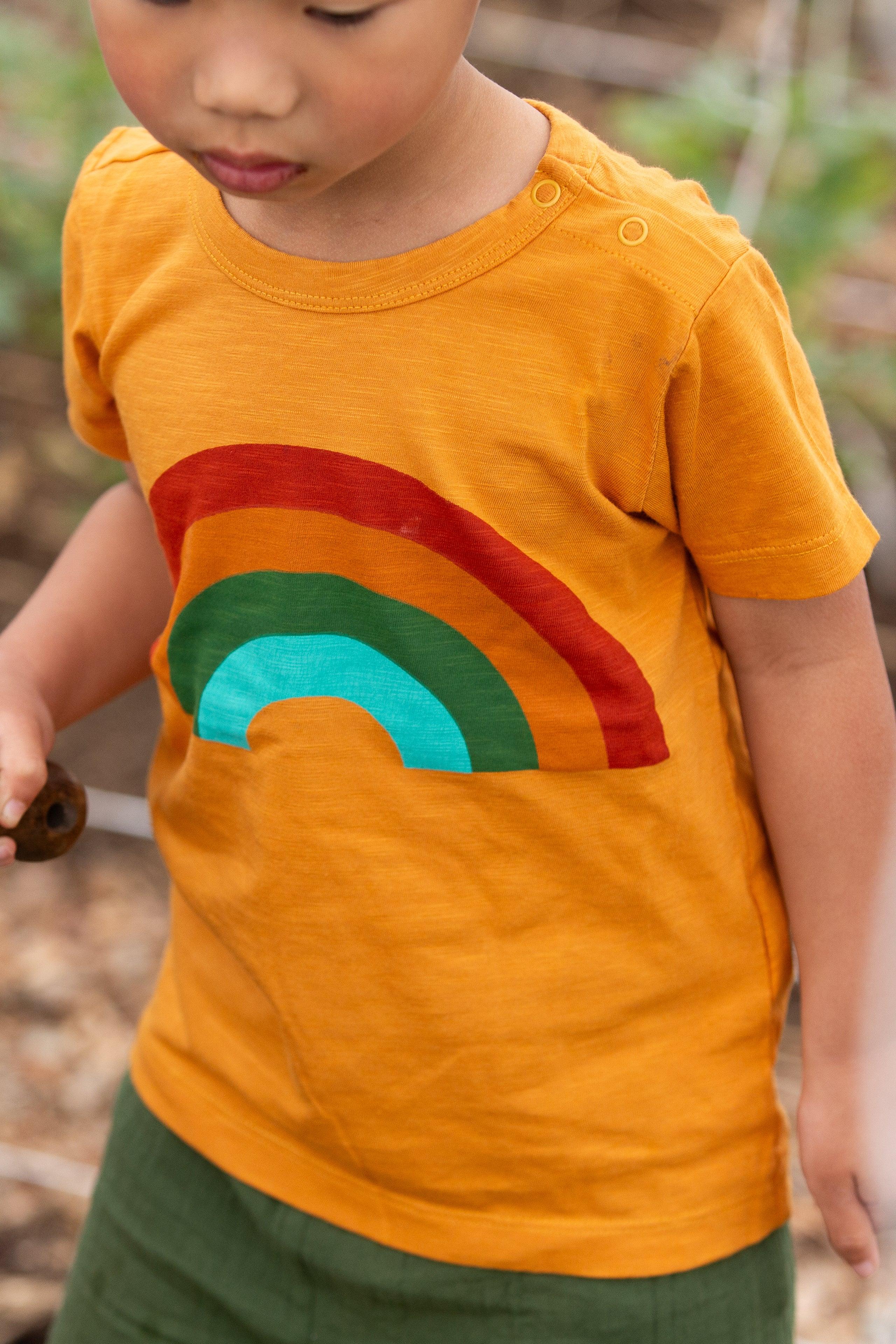 Child wearing an orange shirt with a rainbow design, holding a tool outdoors.