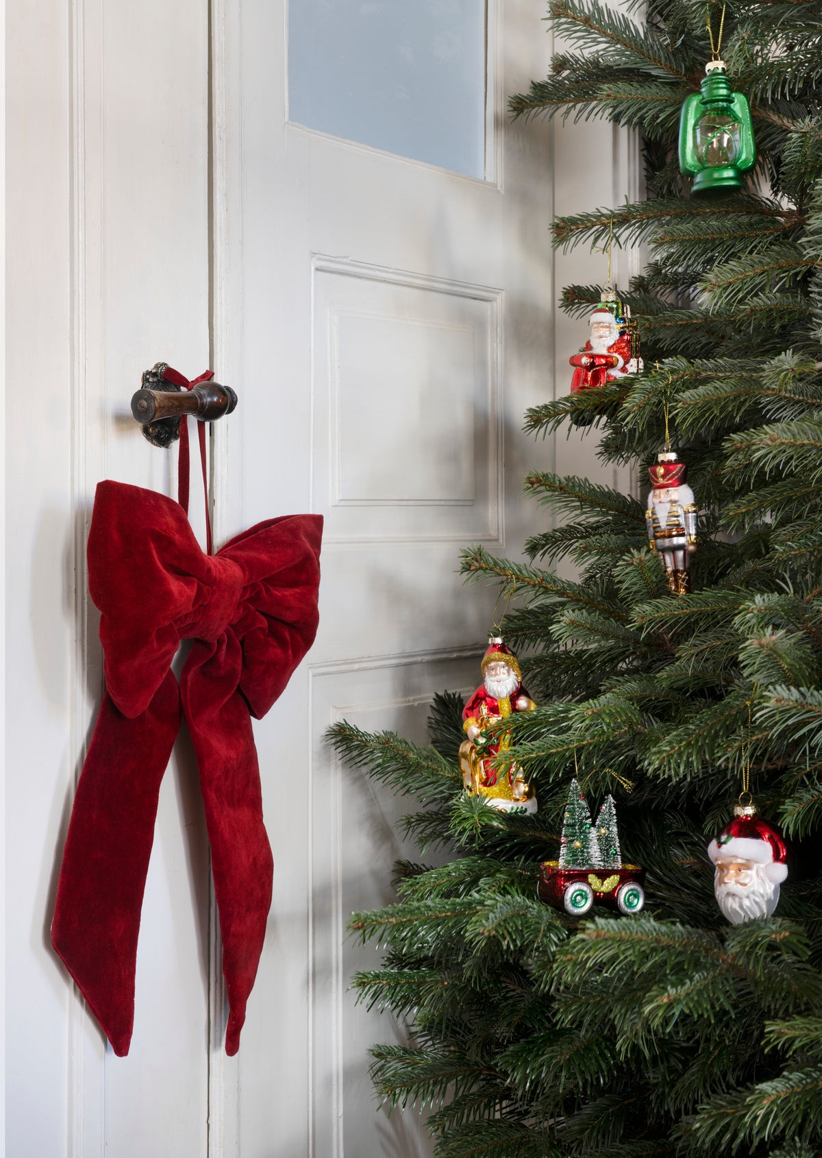 A Christmas tree decorated with various glass Christmas ornaments next to a white door with a large red velvet bow.