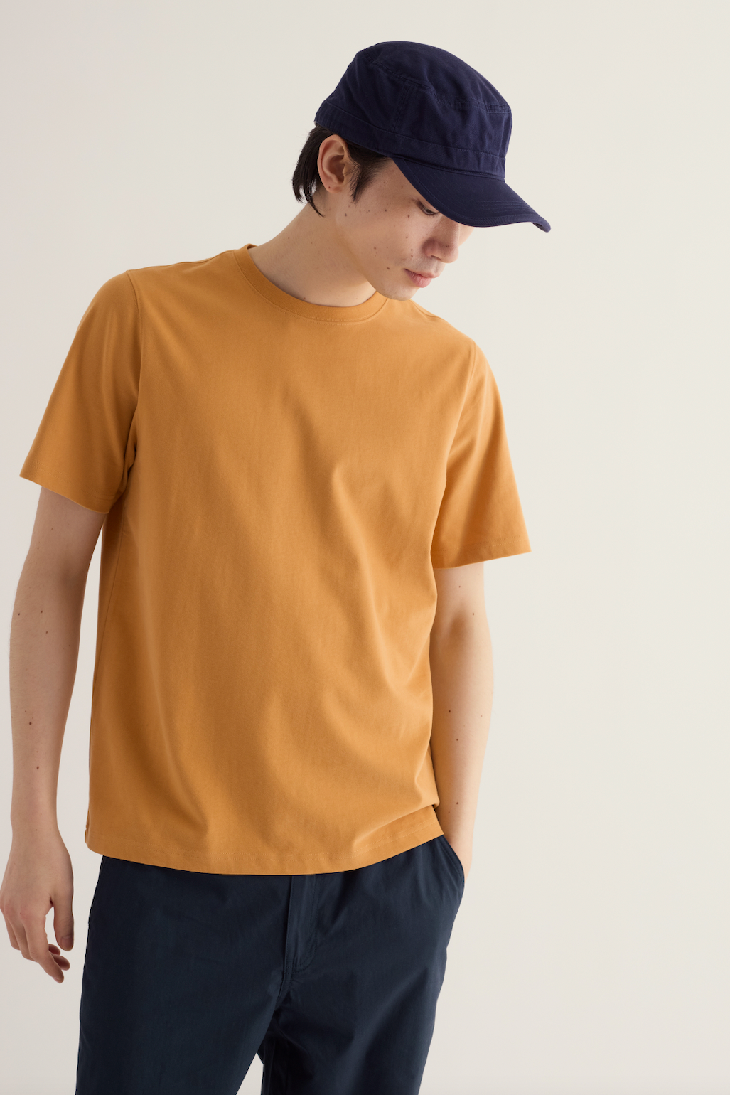 A man wearing a mustard organic cotton mens t-shirt and a dark blue baseball cap looking down.