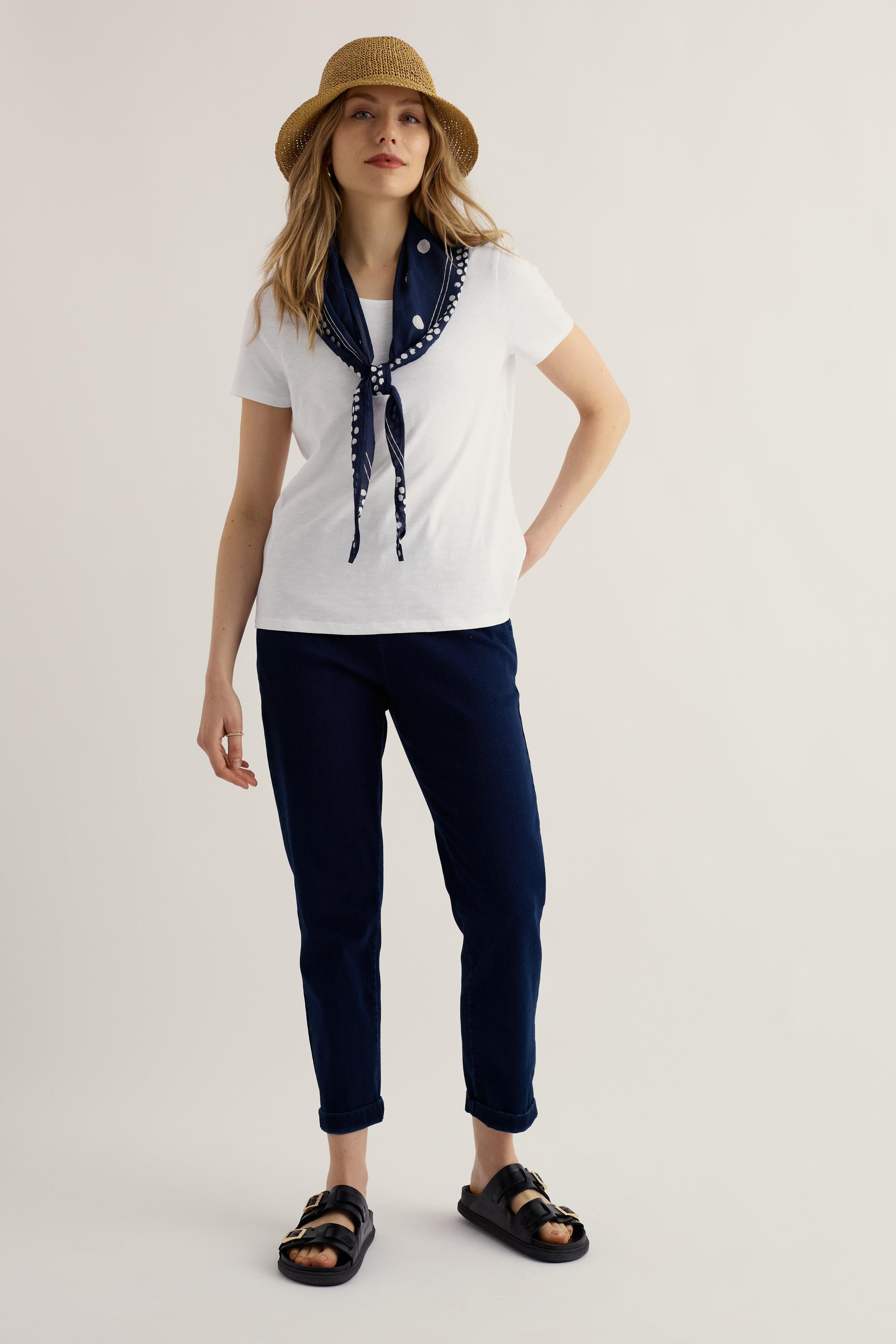 Woman wearing a white organic cotton t-shirt with a navy scarf, navy pants, and a straw hat on a light background